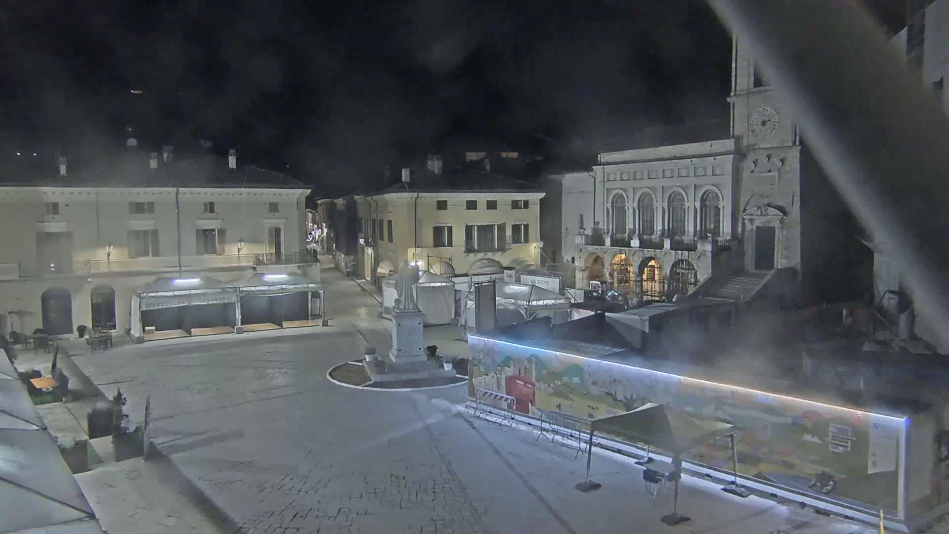 A nighttime view of a town square with several buildings, temporary market stalls, and a statue, under hazy or foggy conditions.