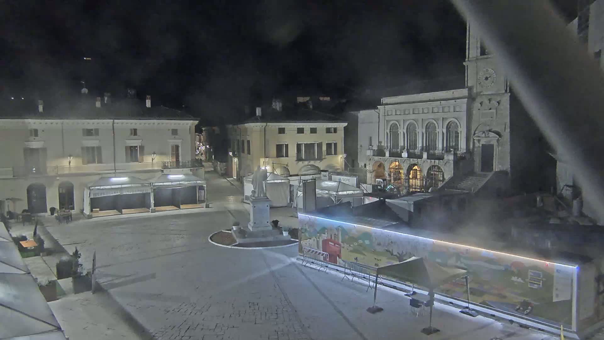 A nighttime view of a town square with several temporary market stalls, a statue, and buildings, under hazy or foggy conditions.