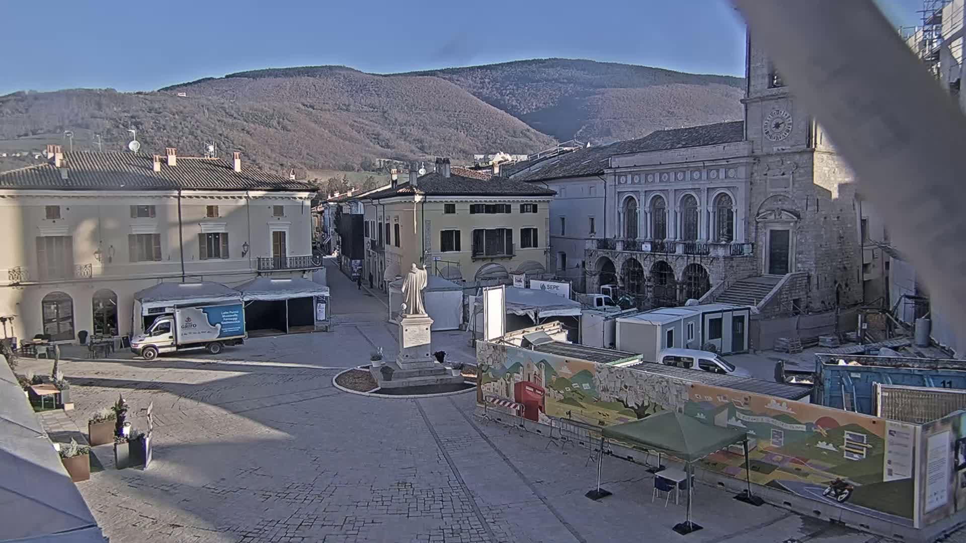 A town square with several buildings, a statue, temporary structures, and construction materials is visible under a clear, sunny sky with mountains in the background.