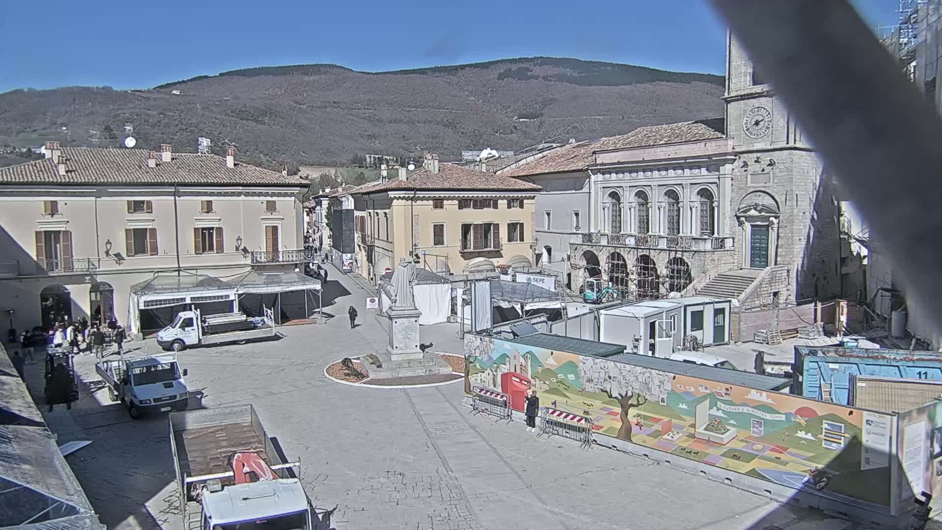 A town square under a sunny sky, surrounded by historic buildings, with construction equipment and temporary structures in the center.