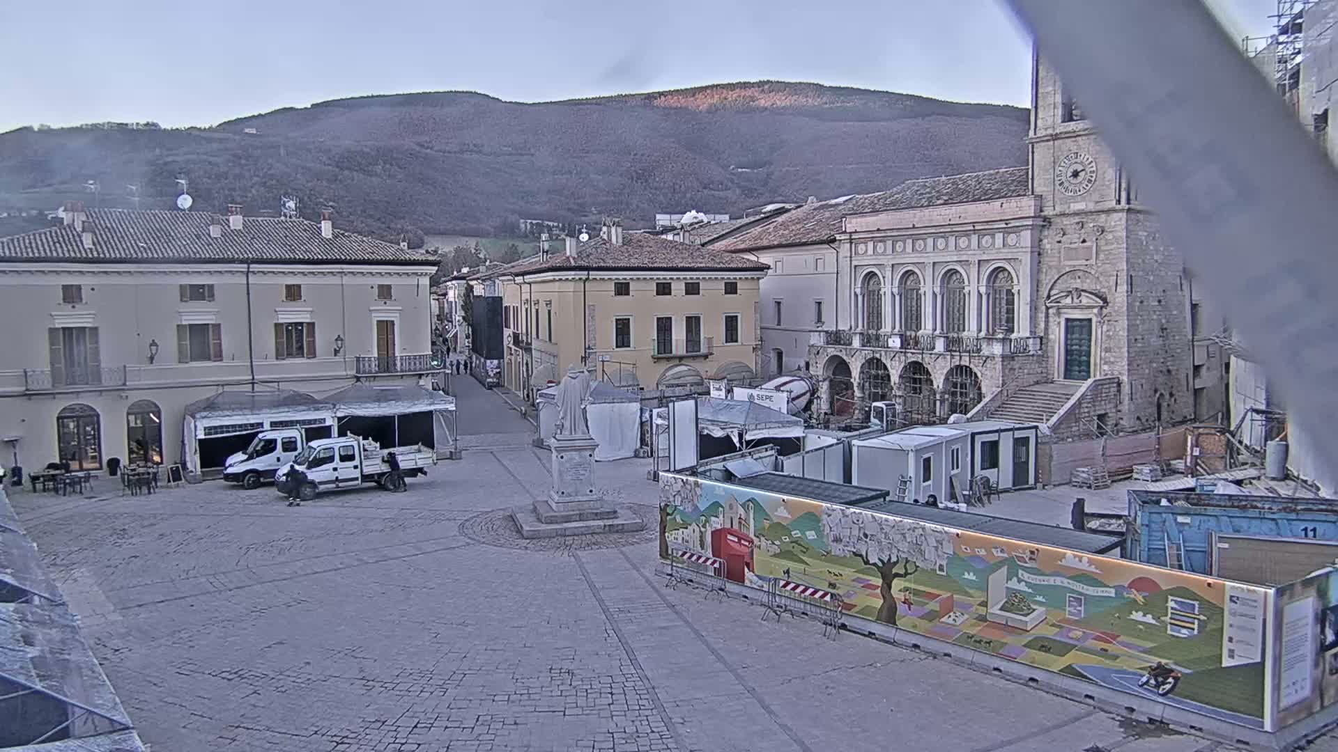 A town square with several buildings, including a large stone structure, is under construction with temporary structures and vehicles present under a clear, pale sky.