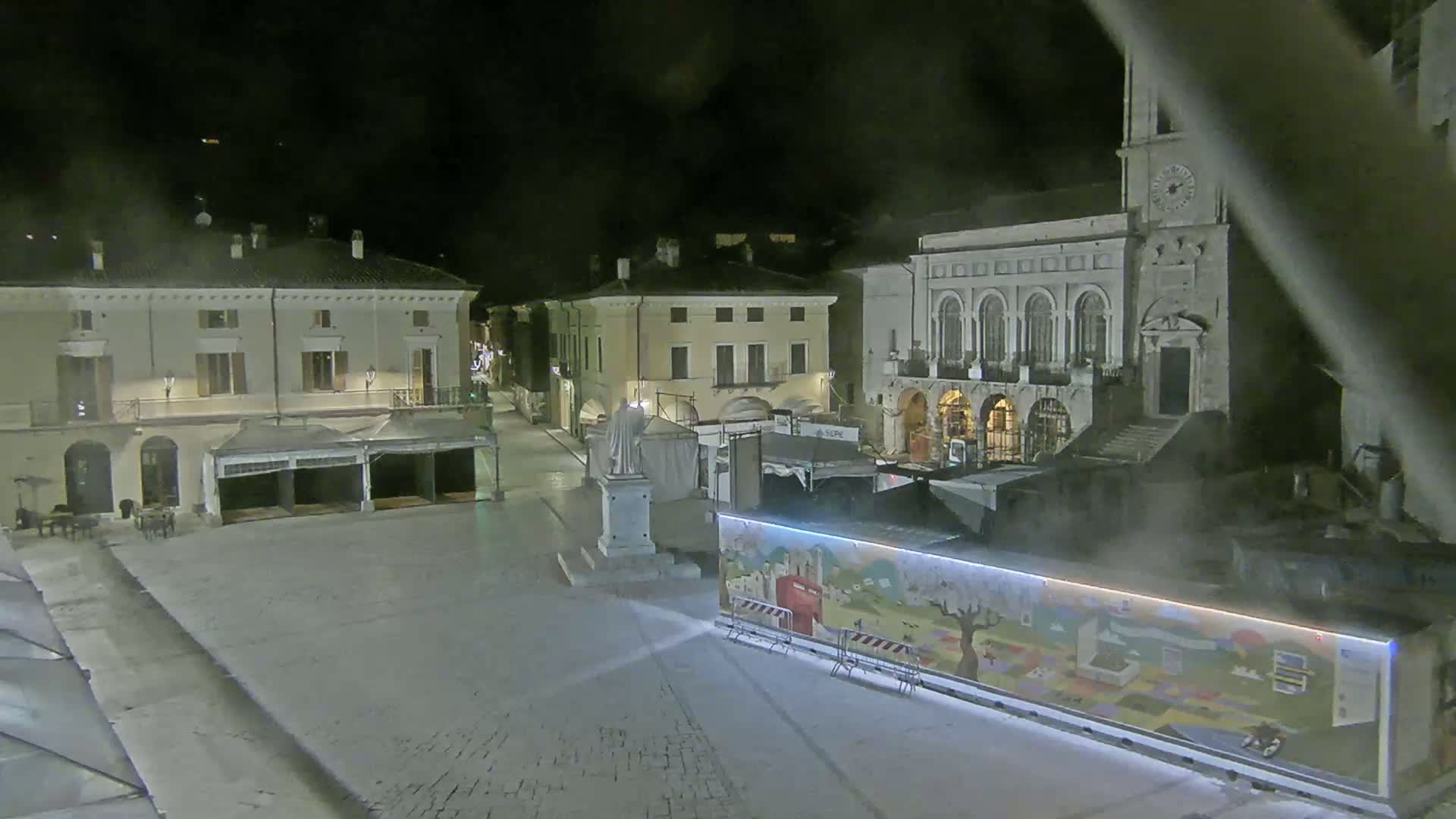 A nighttime view of a town square with several buildings, a statue, and temporary market stalls under a dark, possibly cloudy sky.