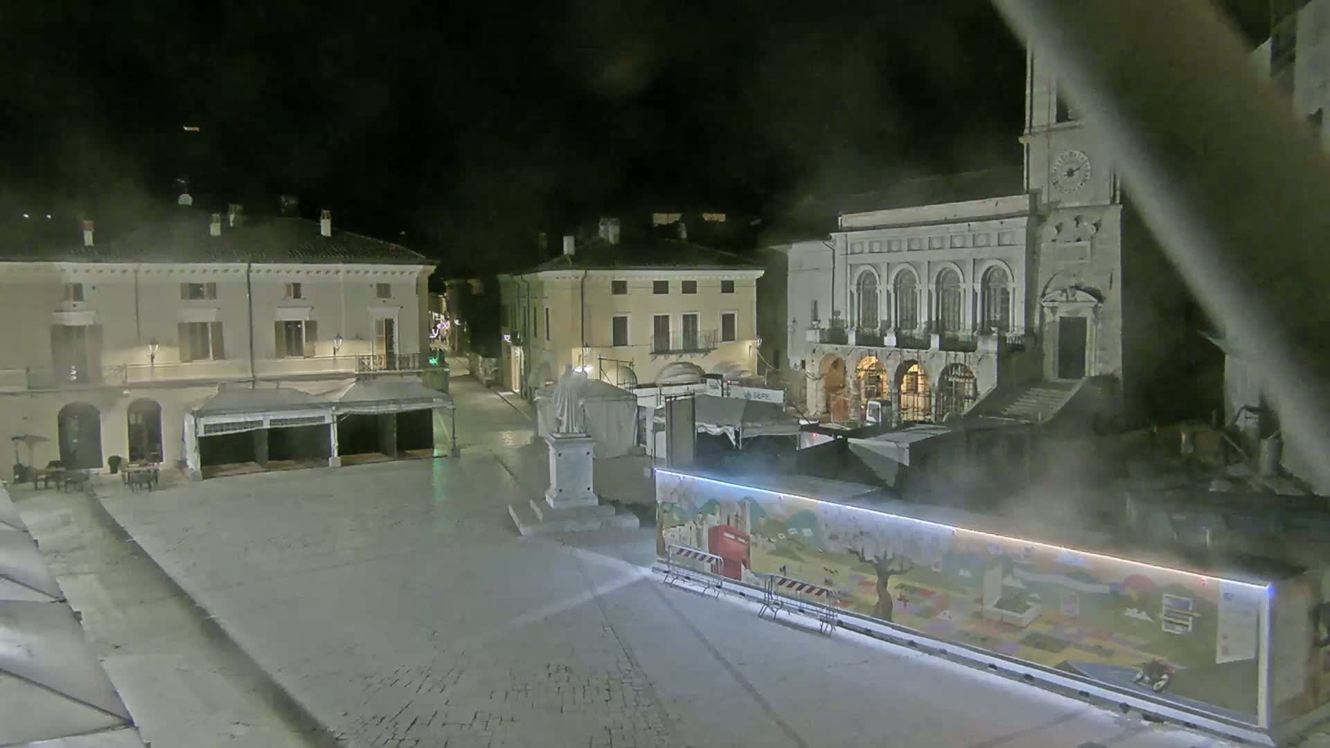 A nighttime view of a town square with several buildings, including a large stone structure, and temporary market stalls or structures, under hazy or foggy conditions.