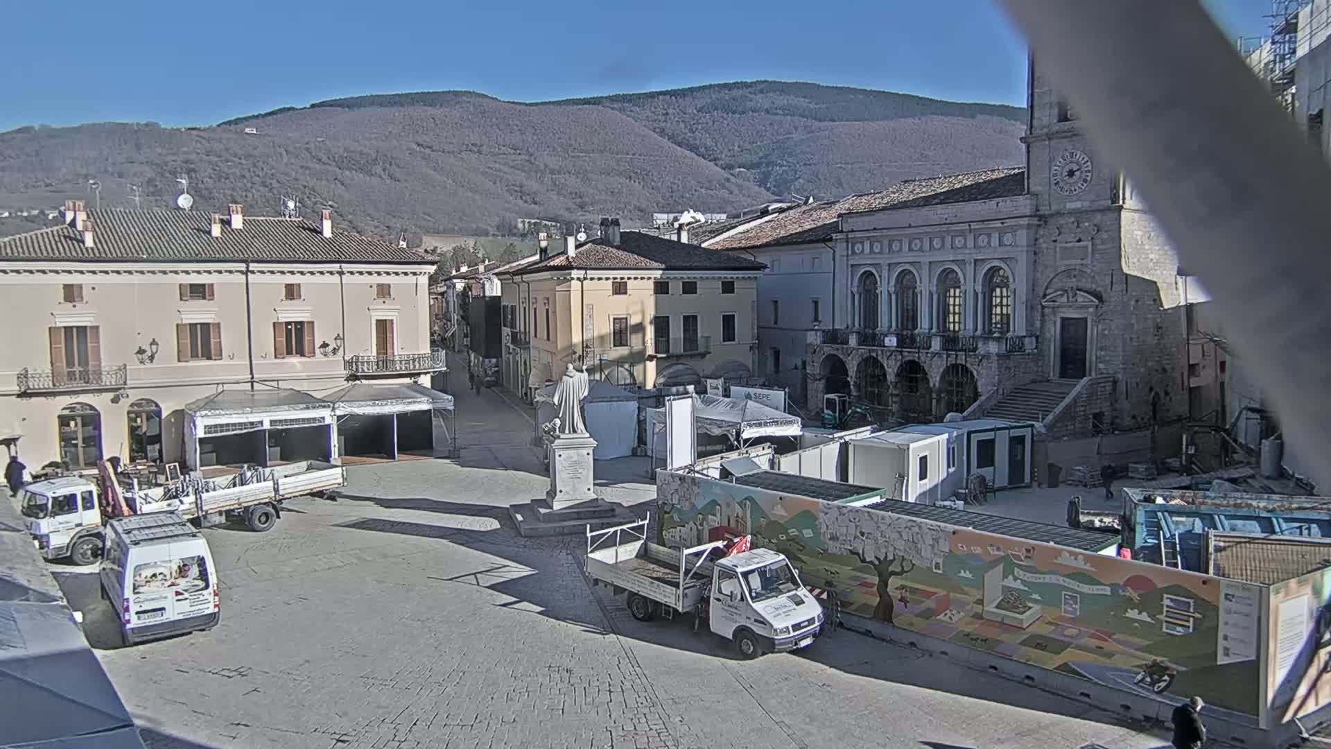 A town square in a mountainous region under sunny skies features several trucks, temporary structures, and a statue, with buildings lining the perimeter.