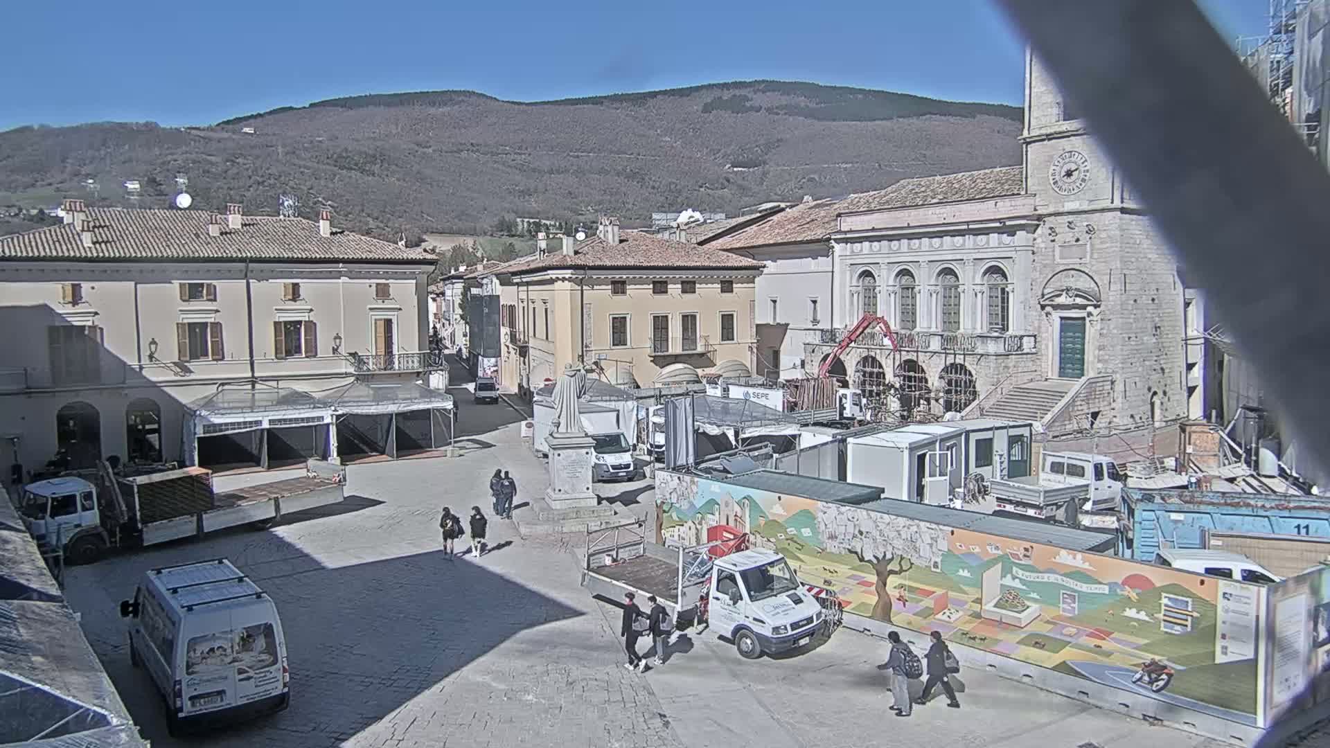 A town square under a clear, sunny sky is filled with construction equipment, temporary structures, and a few pedestrians, with a backdrop of mountains.