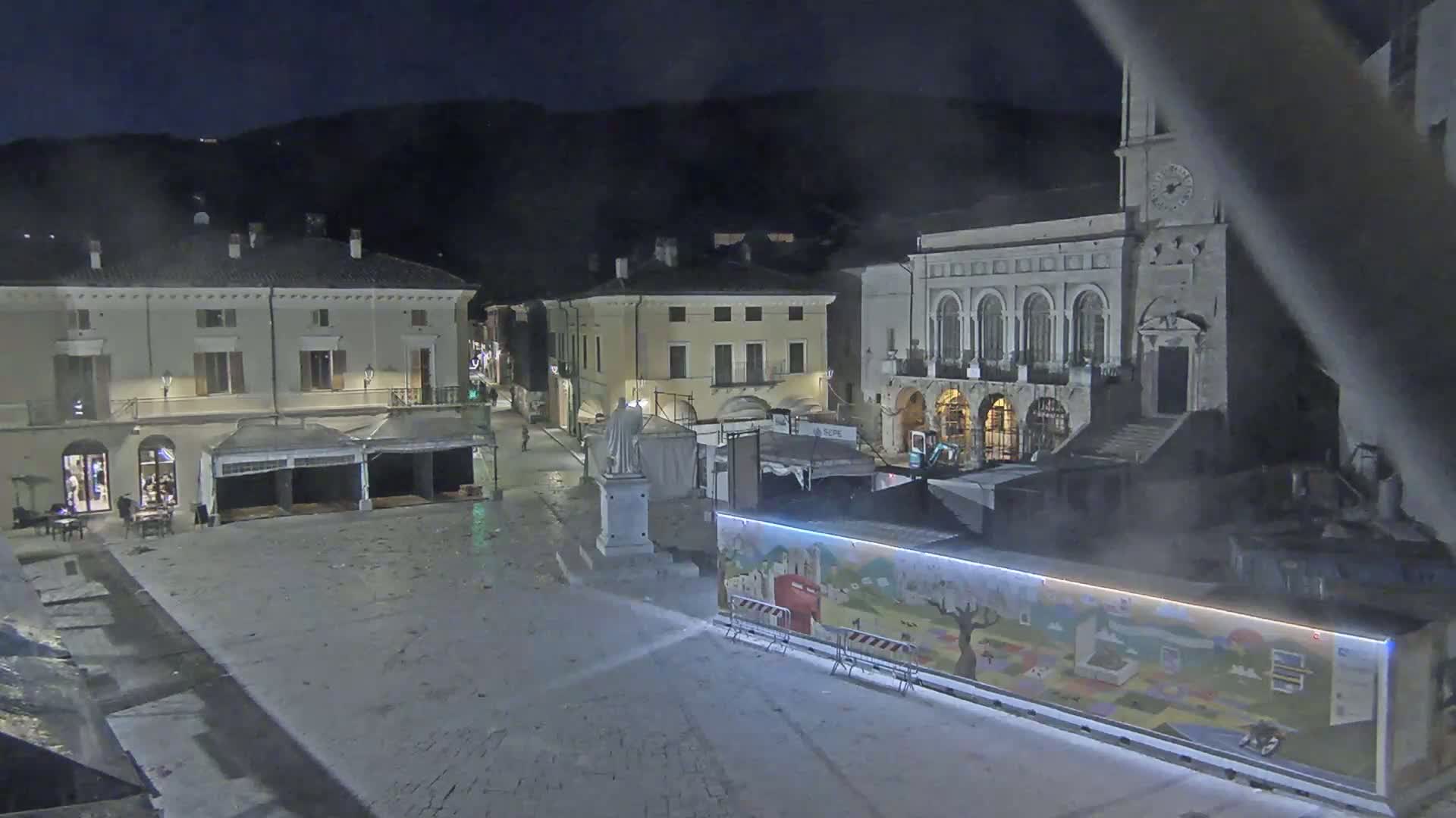 A nighttime view of a town square with several buildings, including a large stone structure, and a brightly-lit mural on a temporary wall.