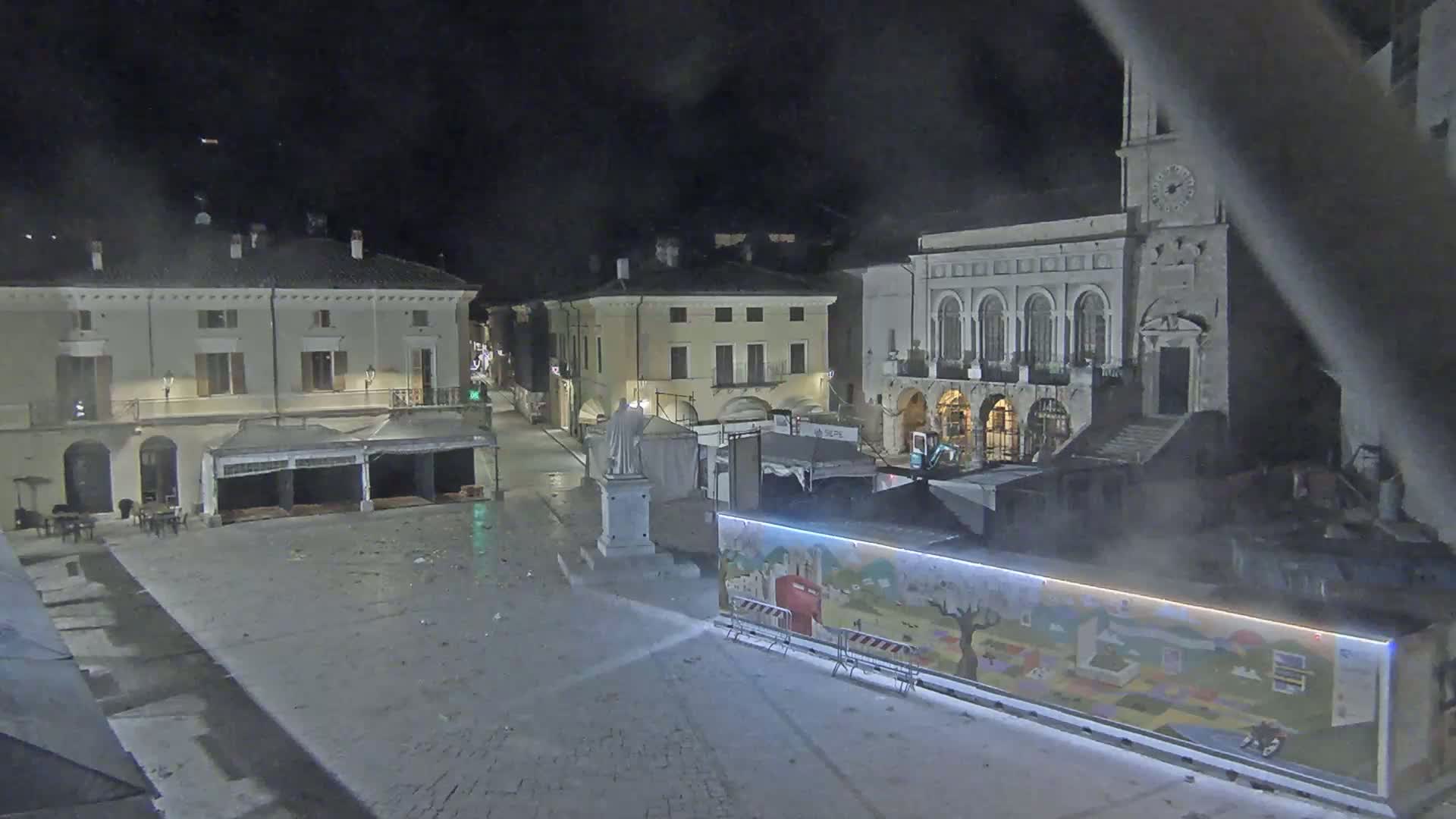 A nighttime view of a town square with several buildings, temporary structures, and a statue, under dark conditions.