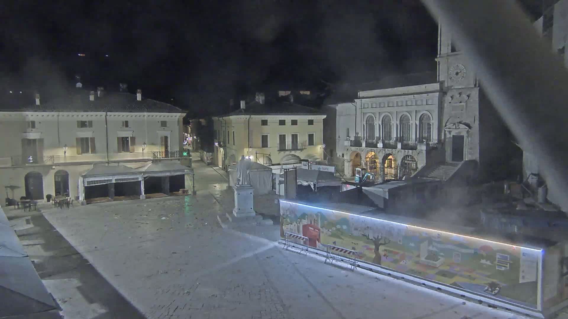 A nighttime view of a town square with several buildings, temporary structures, and a statue, under hazy or foggy conditions.