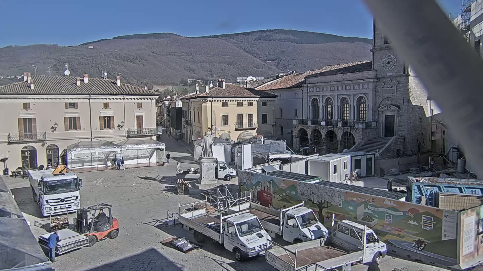 A town square is being prepped for an event on a sunny day, with several trucks, a forklift, and temporary structures in place against a backdrop of buildings and mountains.