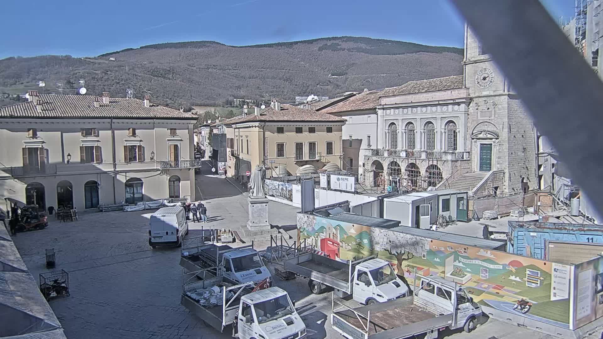 A town square under a sunny sky is partially blocked off by construction materials and several trucks, with several buildings and a mountain in the background.