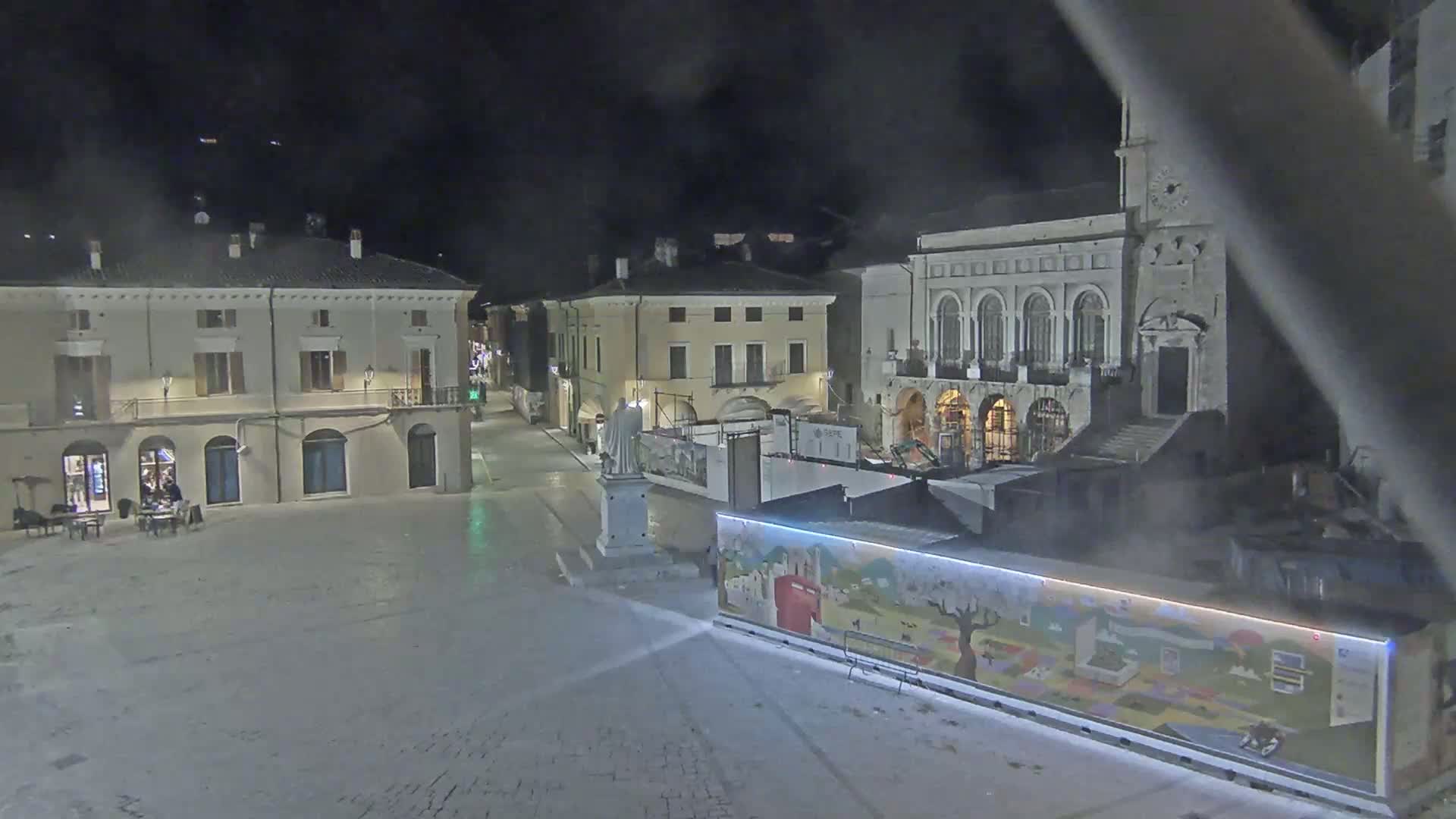 A mostly empty town square at night is illuminated, showing several buildings, a statue on a pedestal, and a colorful structure in the foreground.