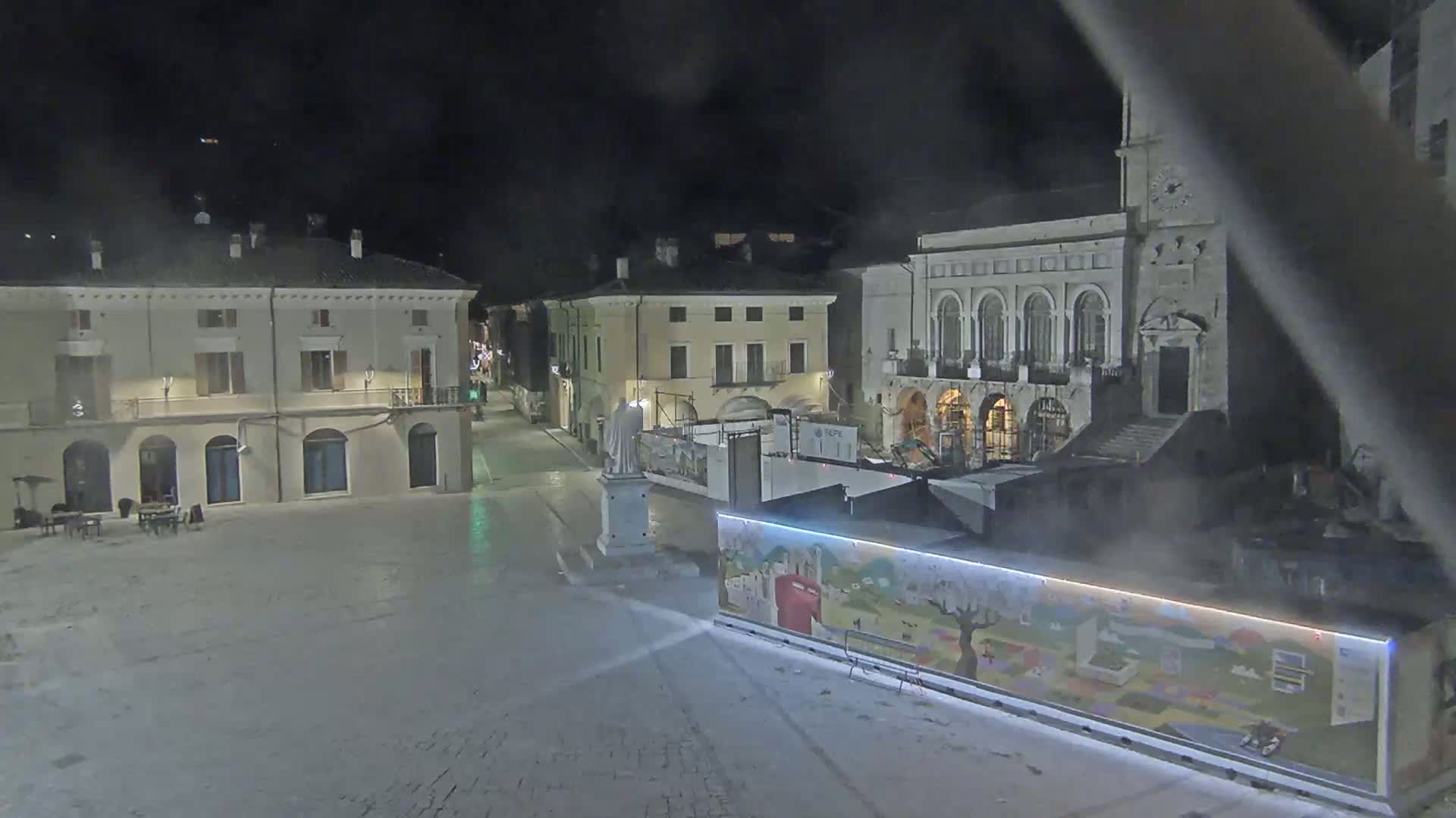 A mostly empty town square at night is shown, with several buildings surrounding it, under hazy conditions.