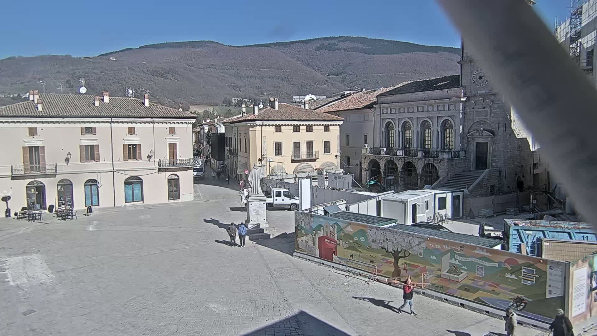 A town square with several buildings, a statue, and construction underway, under a clear sunny sky and mountainous backdrop.