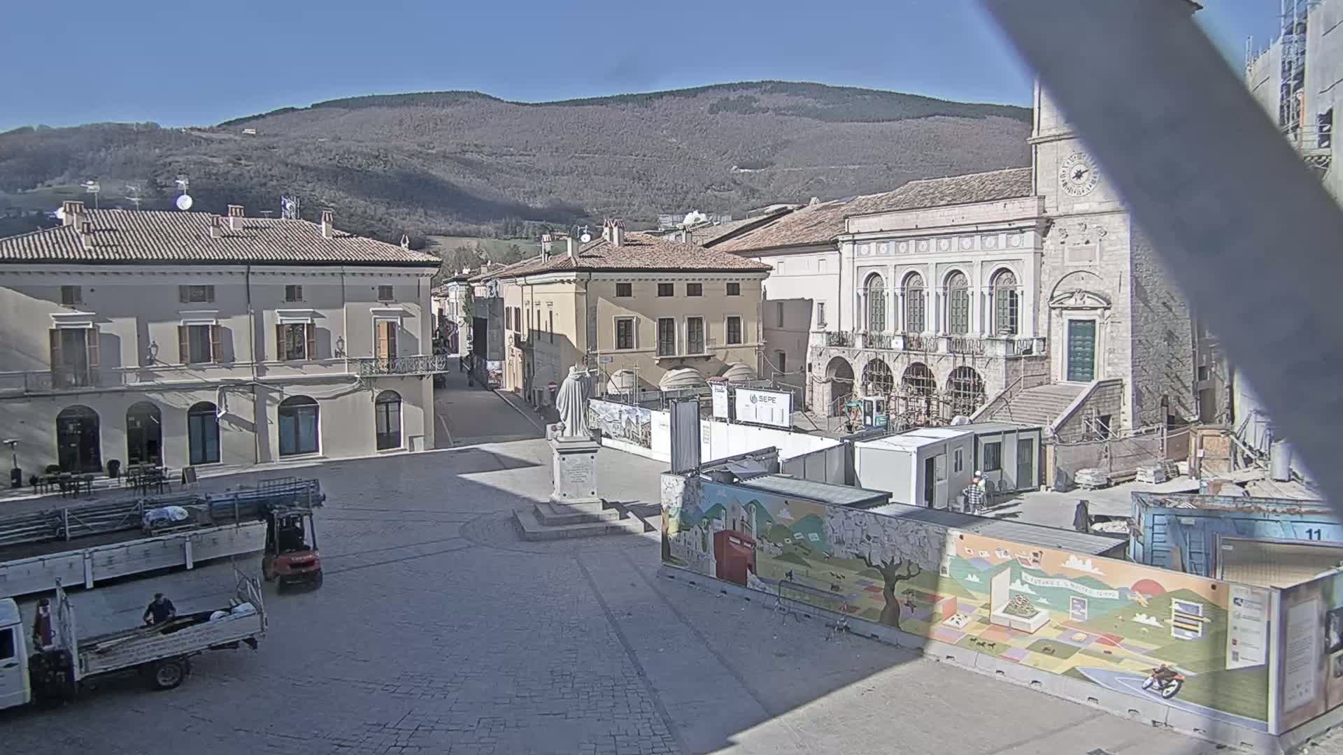 A town square under a sunny sky is partially obscured by construction, featuring several buildings, a statue, and trucks.