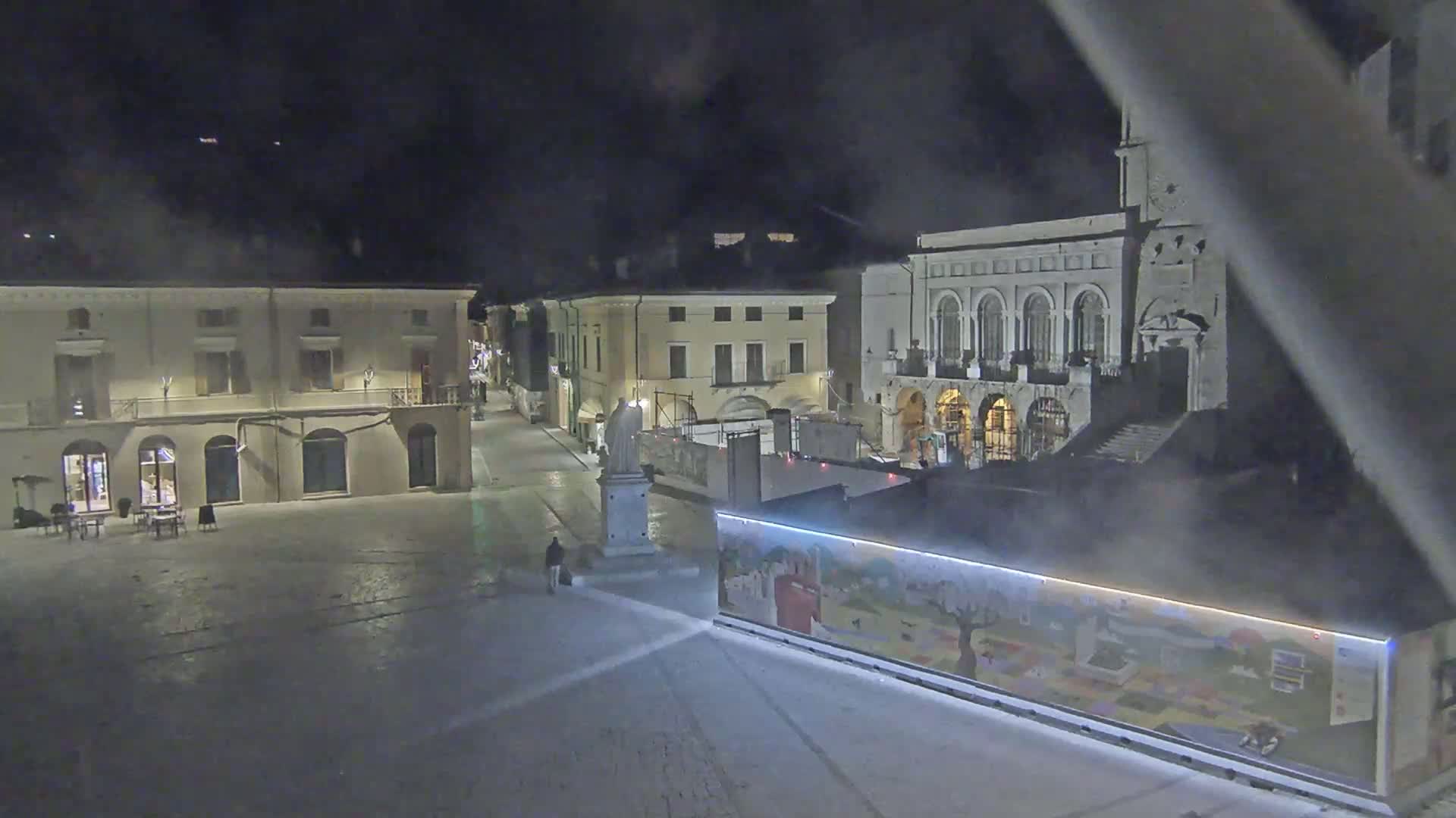 A nighttime view of a town square with several buildings, one of which has a clock tower, under hazy conditions.