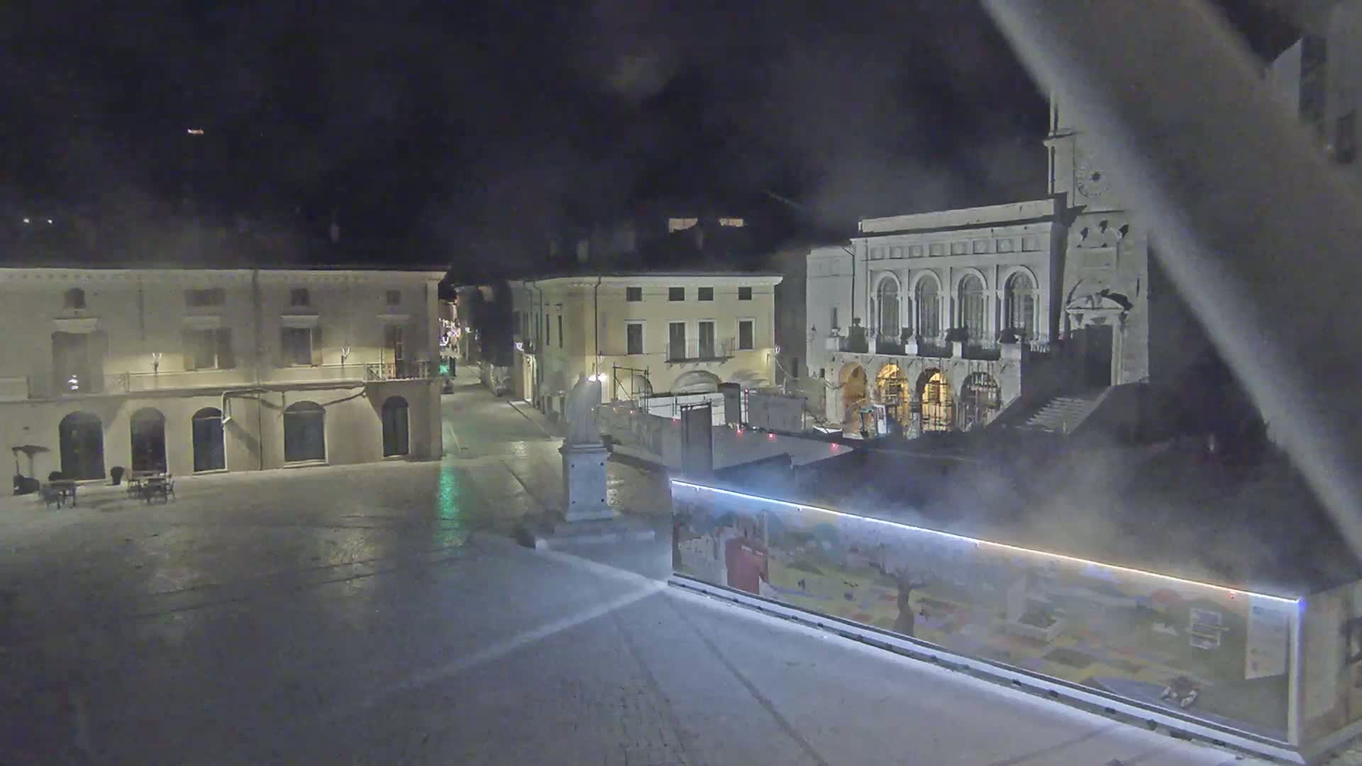 A nighttime, hazy view of a town square with several buildings, including a large one with arched windows and a clock tower, partially obscured by what appears to be construction.