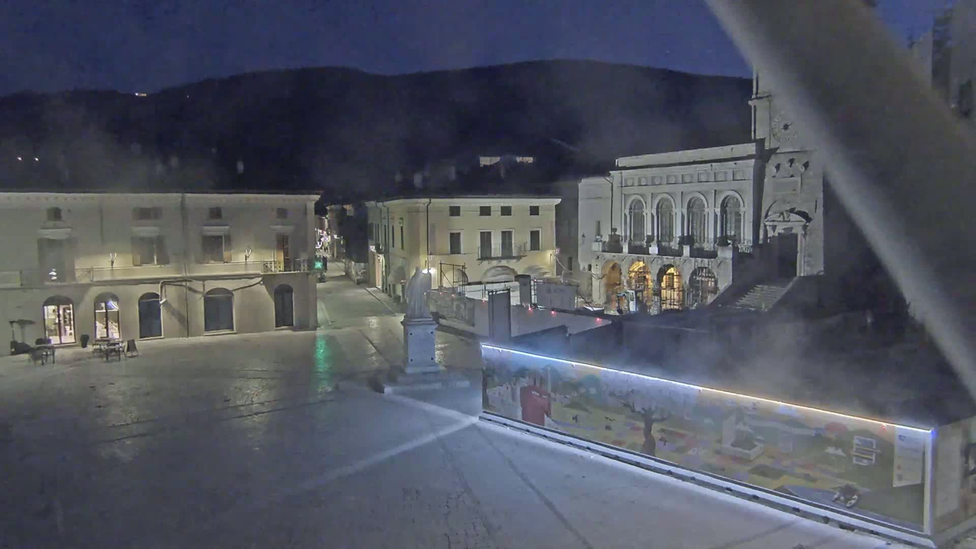 A nighttime view of a town square with several buildings, including a large one with arched windows, under a dark, possibly foggy sky.
