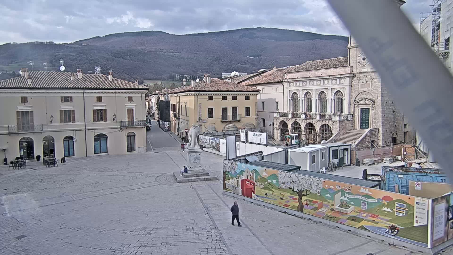 A town square under a cloudy sky, with several buildings surrounding it, one of which is under construction and partially obscured by a colorful mural-covered construction barrier.