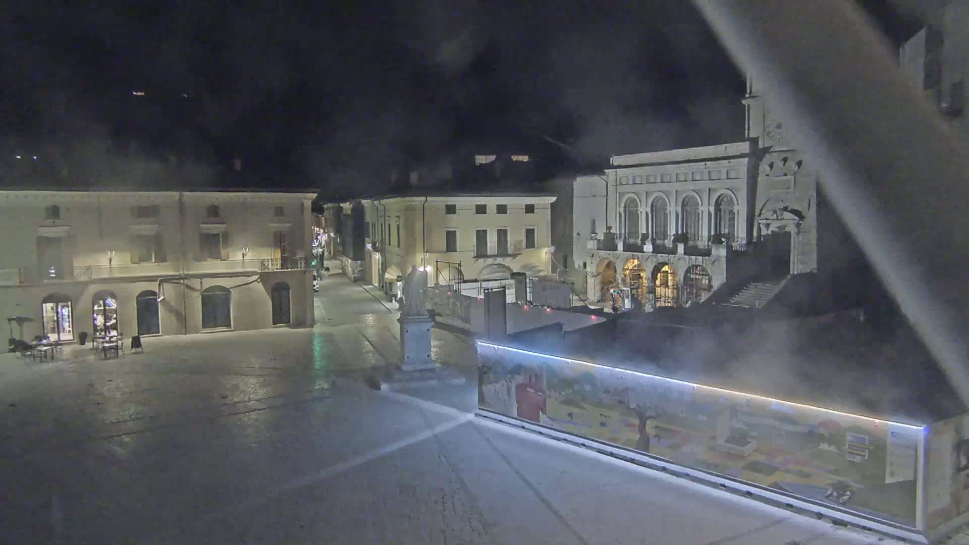 A nighttime view of a mostly empty town square with several buildings, one of which has a clock tower, under a hazy sky.