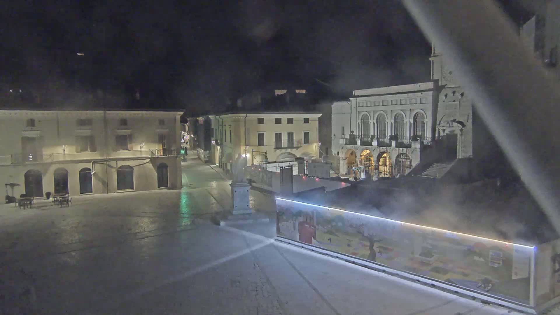 A nighttime view of a town square with several buildings, including a large building with arched windows, under hazy or foggy conditions.