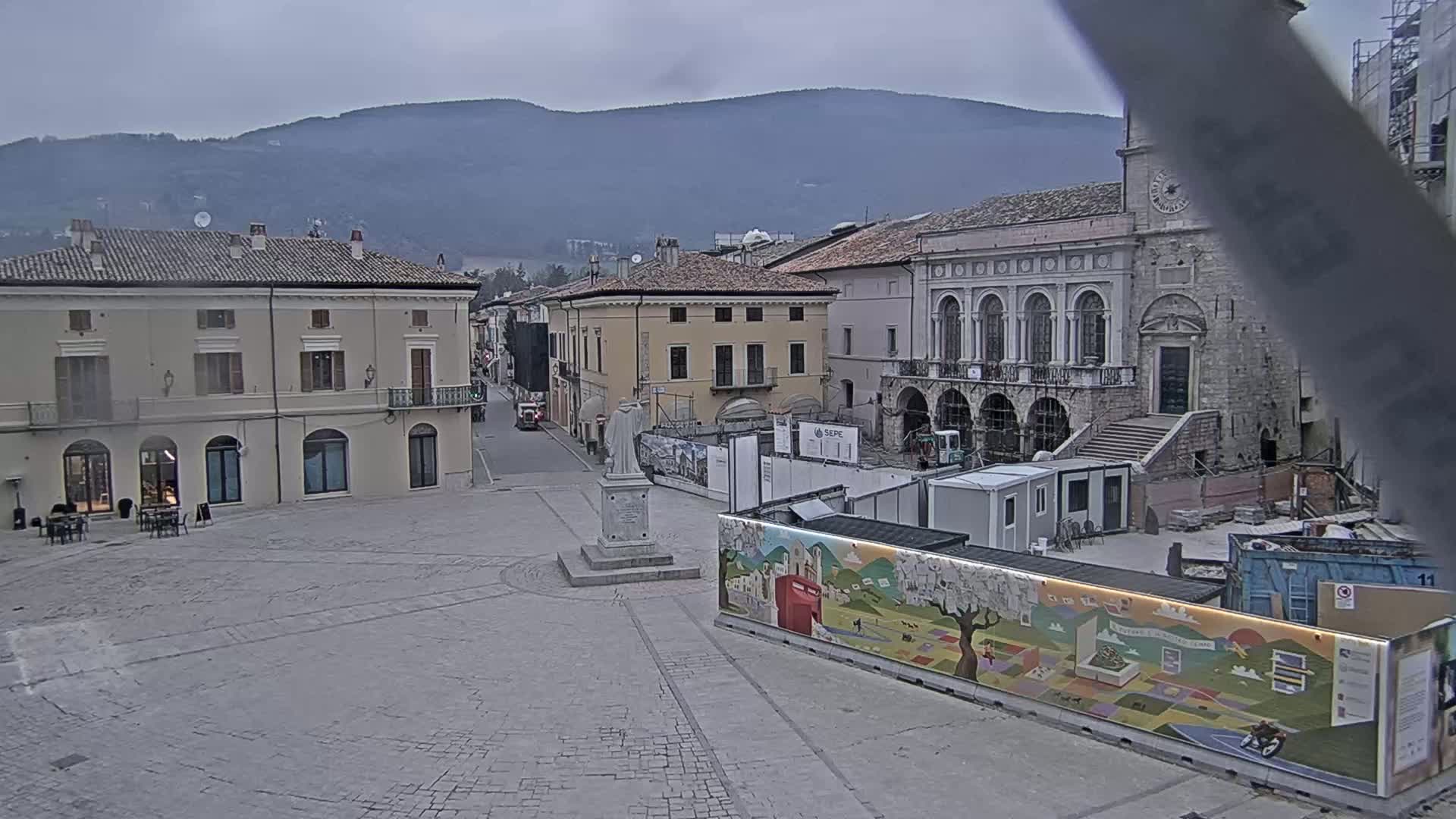 A town square with several buildings, a statue, and construction underway under an overcast sky.