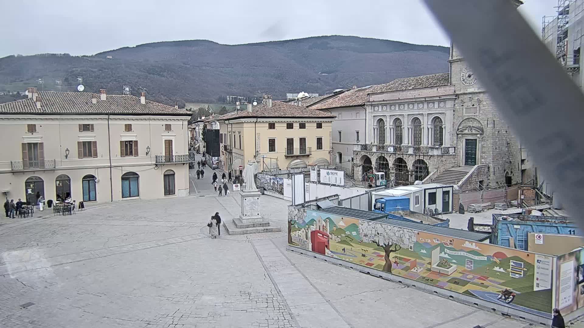 A town square under a cloudy sky shows buildings under construction, with a statue in the center and a few pedestrians.