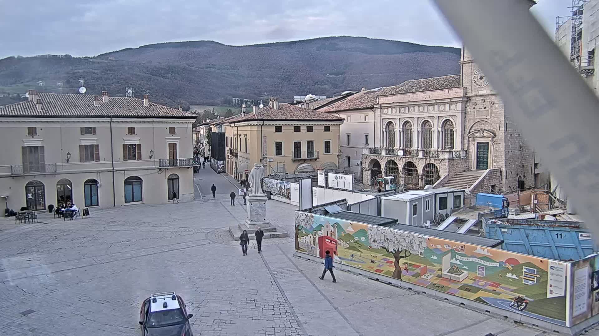 A town square under an overcast sky features several buildings, a statue, a few pedestrians, and construction barriers with colorful artwork, set against a backdrop of distant mountains.