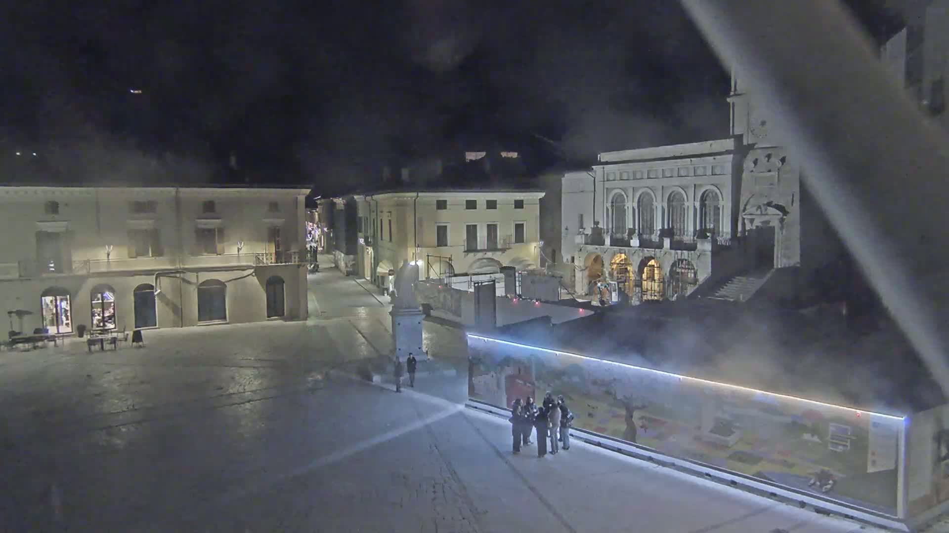 A nighttime view of a town square with several buildings, a statue, and a few people gathered near a large illustrated billboard, under hazy conditions.