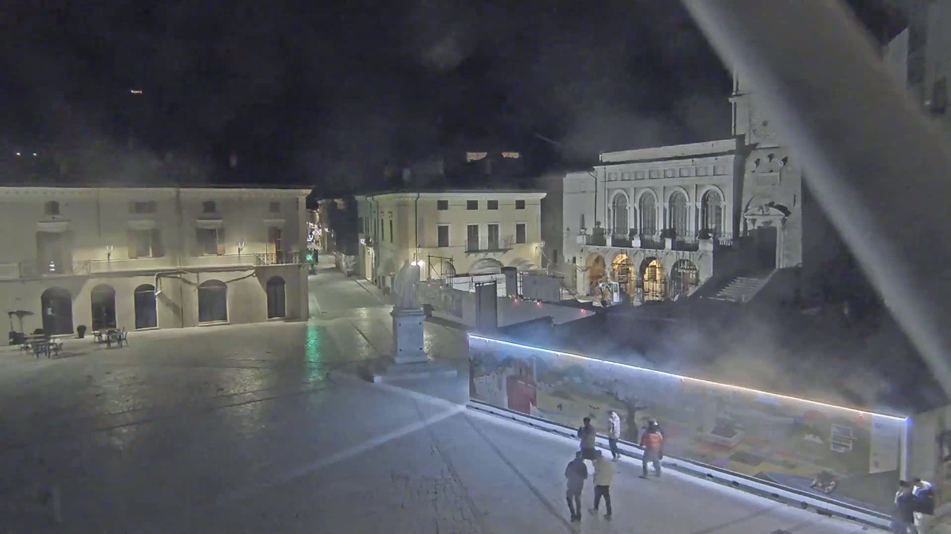 A nighttime view of a town square with several buildings, a statue, and a few people standing near a large, colorful wall, under hazy or foggy conditions.