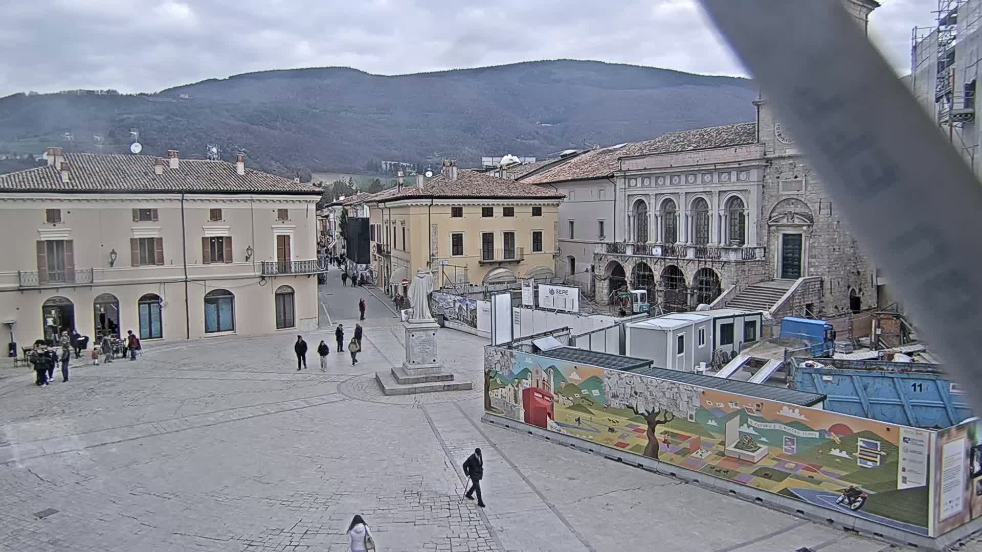 A town square under an overcast sky, with several buildings surrounding it, one of which is under construction, and a few people walking around.