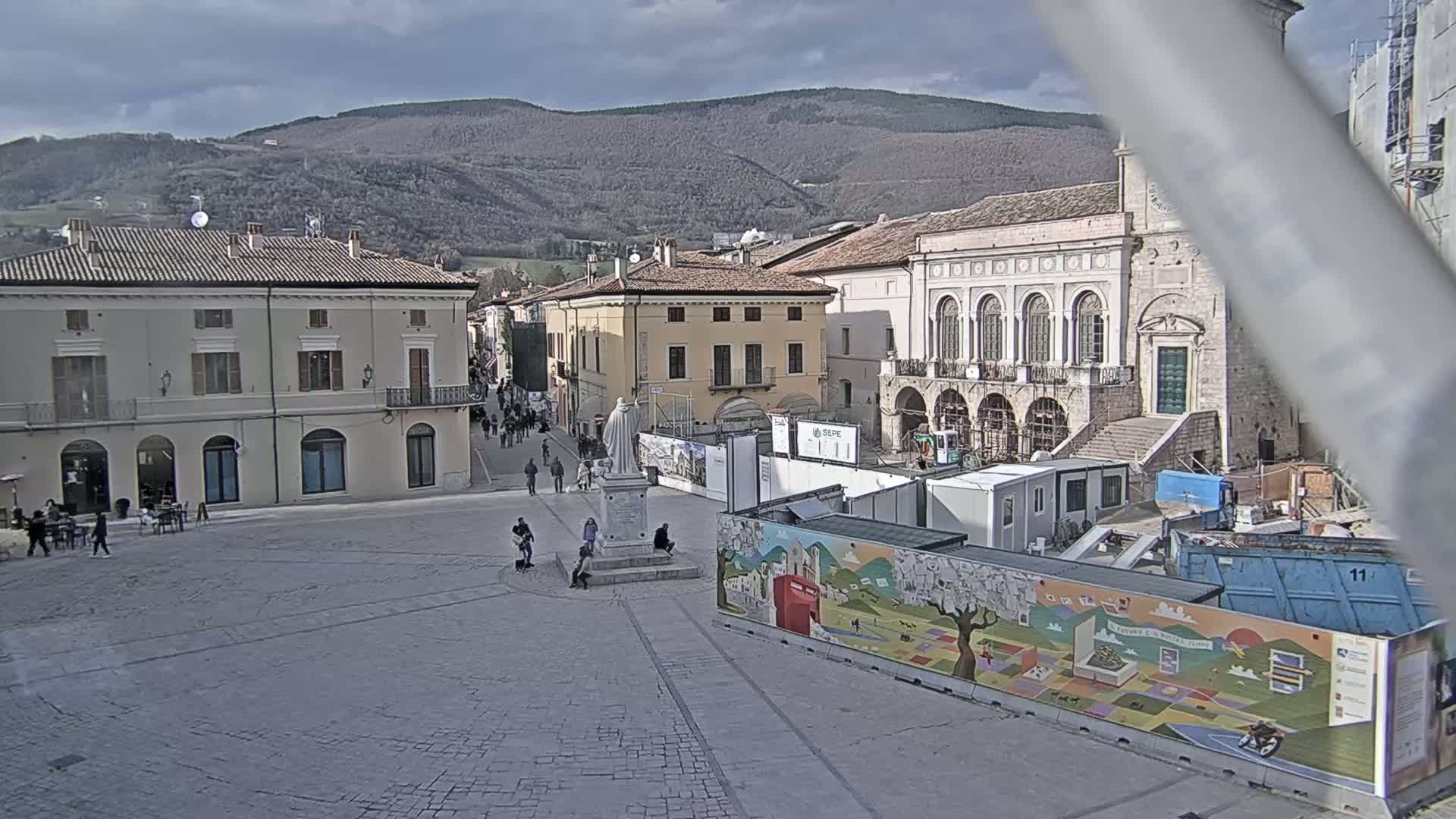 A town square with several buildings, a statue, and some people, under a cloudy sky with mountains in the background, and construction underway in part of the square.