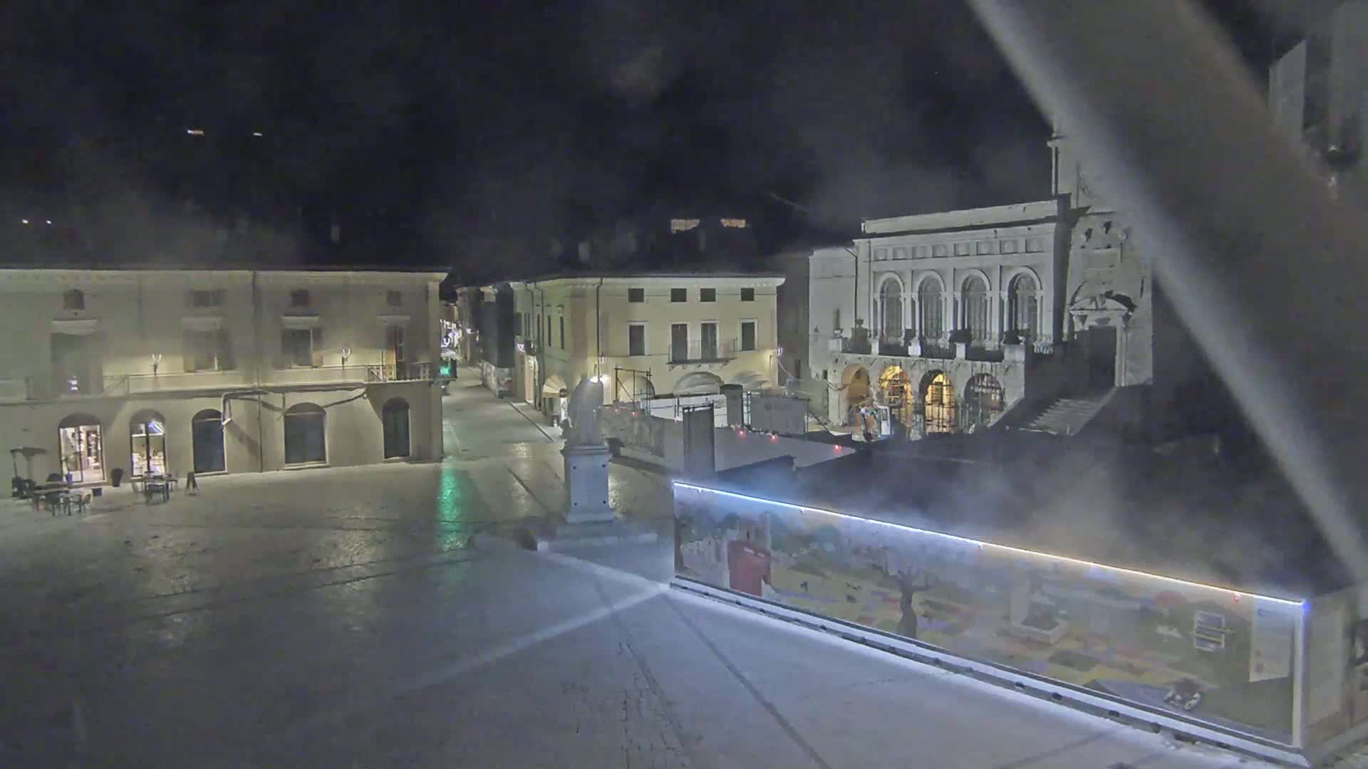 A nighttime view of a town square with several buildings, one of which is ornate, and a large, colorful billboard partially obscuring the view.