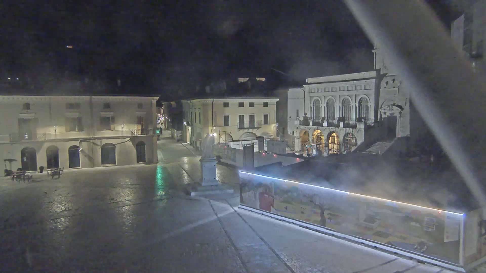 A mostly empty, cobblestone town square at night is lit by ambient light, with several buildings surrounding it; steam is visible rising from something in the square.