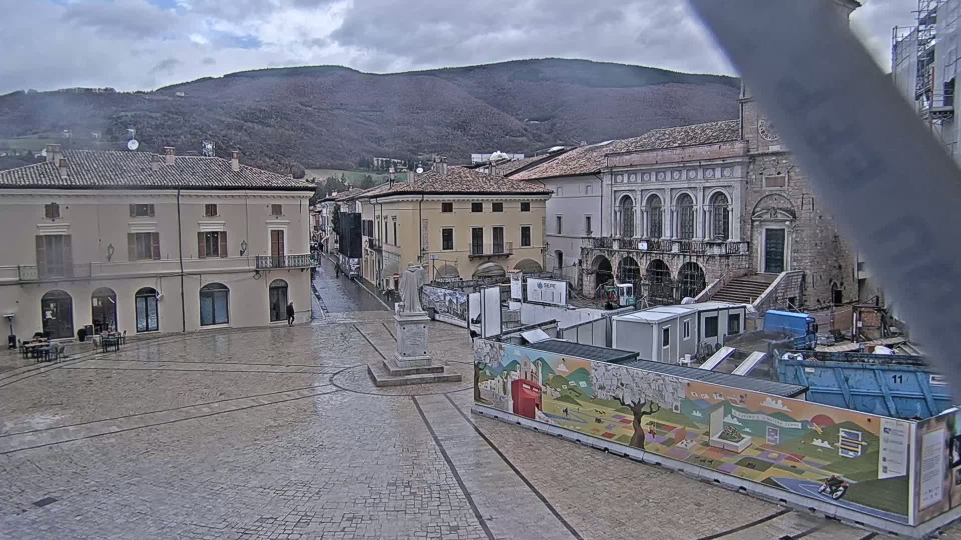 A wet cobblestone town square is surrounded by historic buildings under a cloudy sky, with construction taking place in the center.