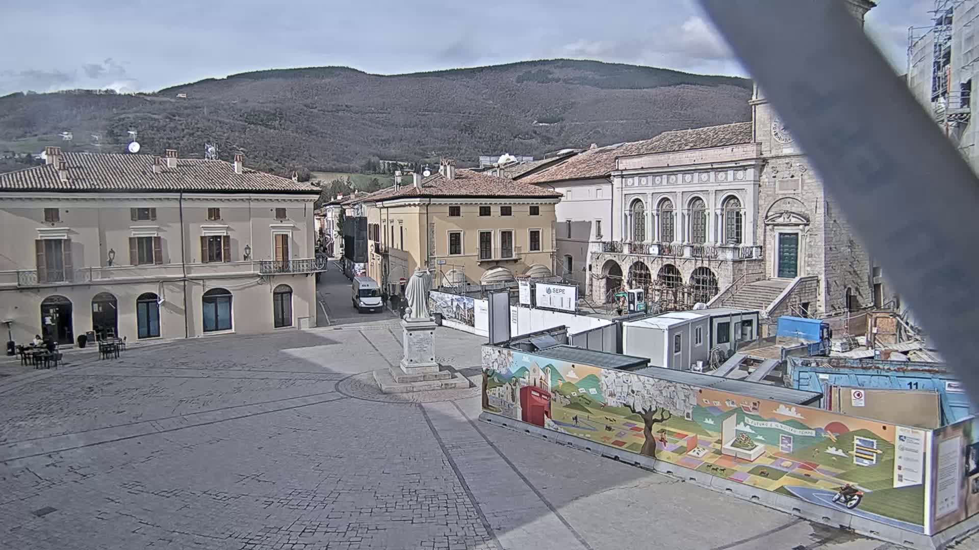 A town square with several buildings, a statue, and construction underway under a mostly clear sky with mountains in the background.