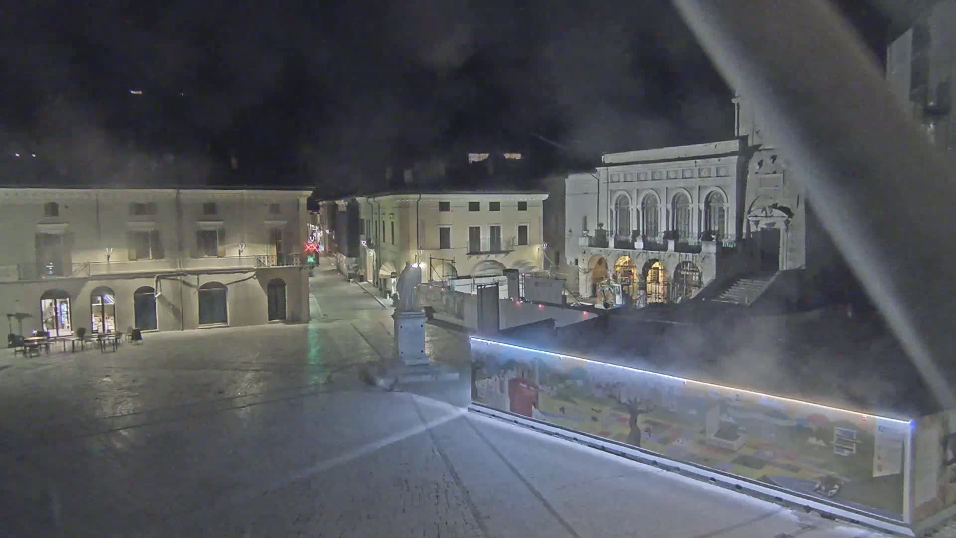 A nighttime view of a snow-covered town square with several buildings, a statue, and a construction-like barrier with a painted mural.