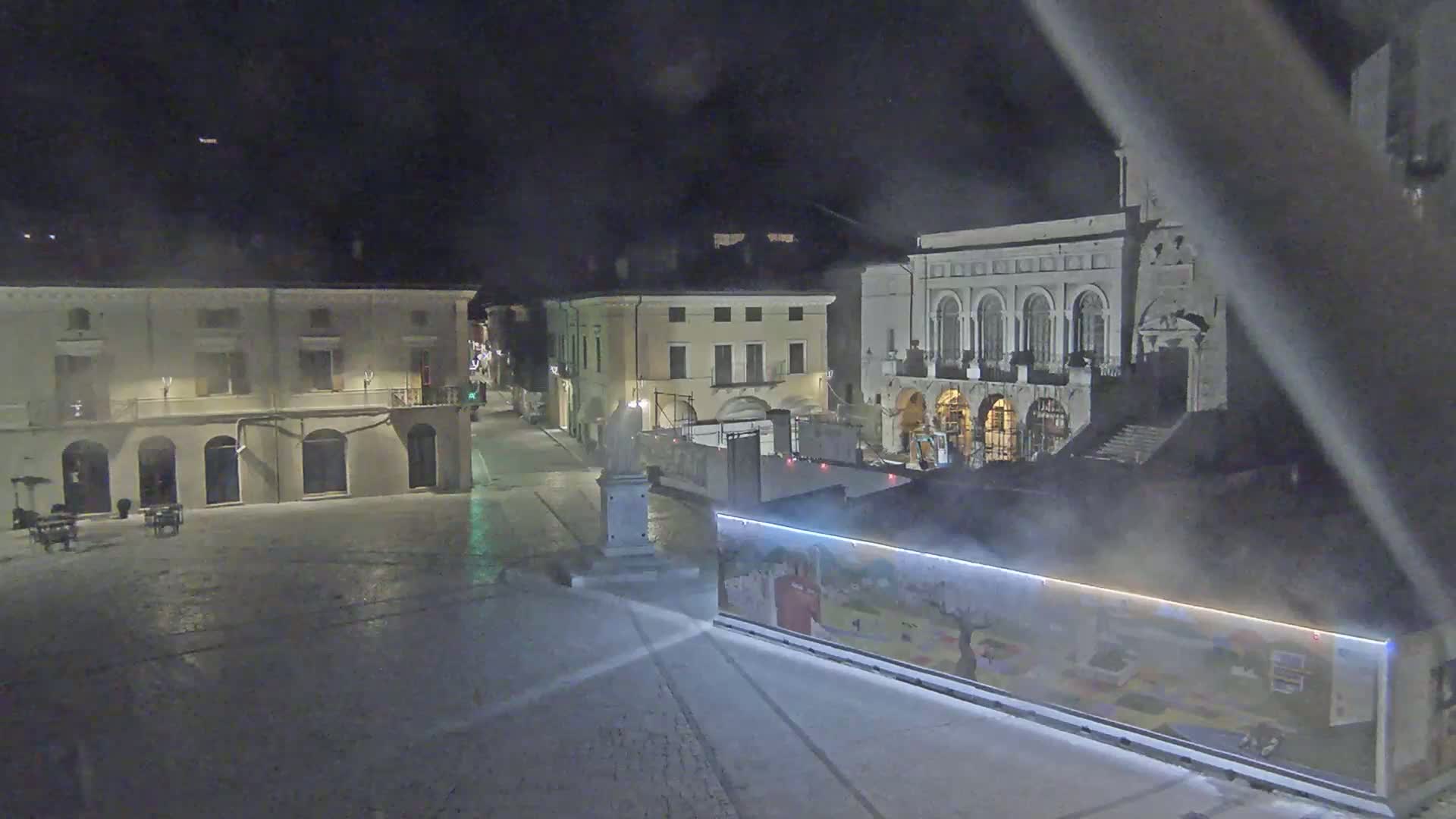 A mostly empty town square at night, with several buildings surrounding it, is lightly covered in a thin layer of mist or fog.