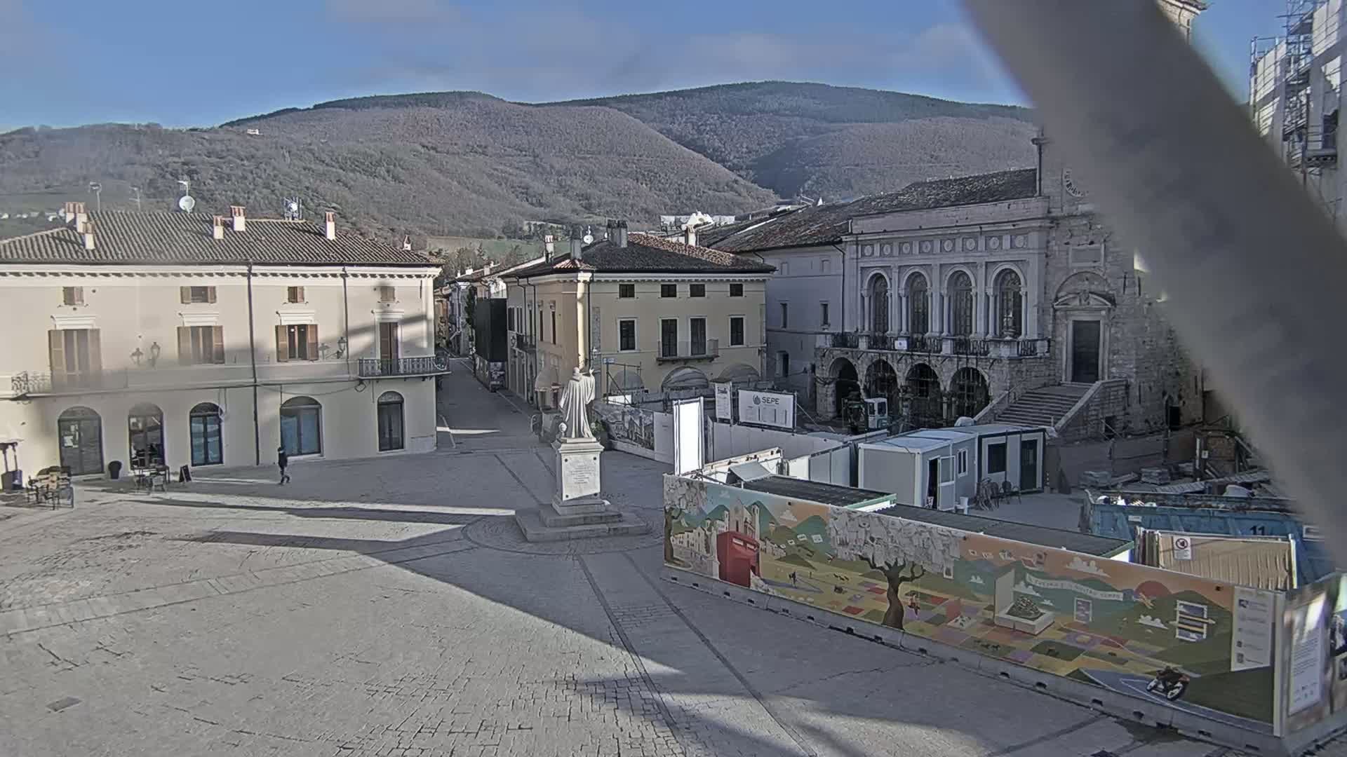 A town square with several buildings, a statue, and construction underway, all under a clear sunny sky with mountains in the background.