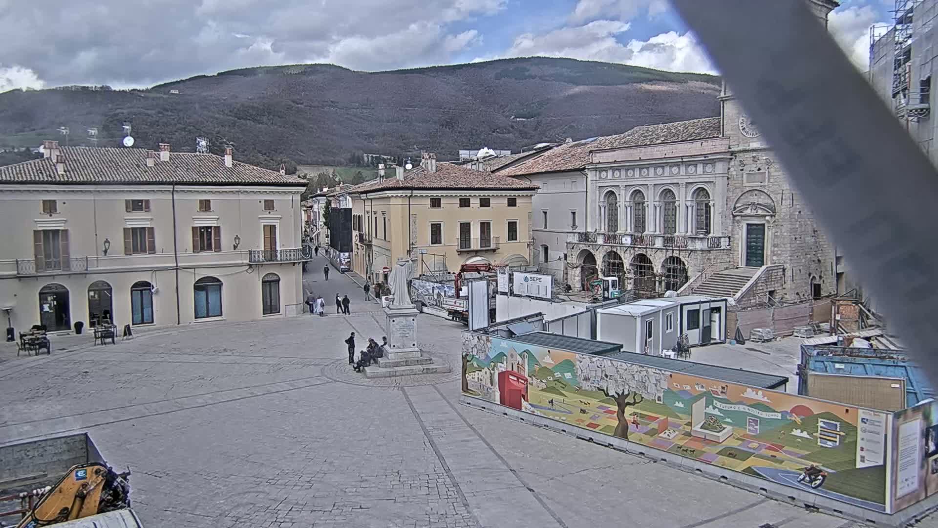 A town square under a cloudy sky, with several buildings surrounding it, some of which are under construction, and a monument in the center.
