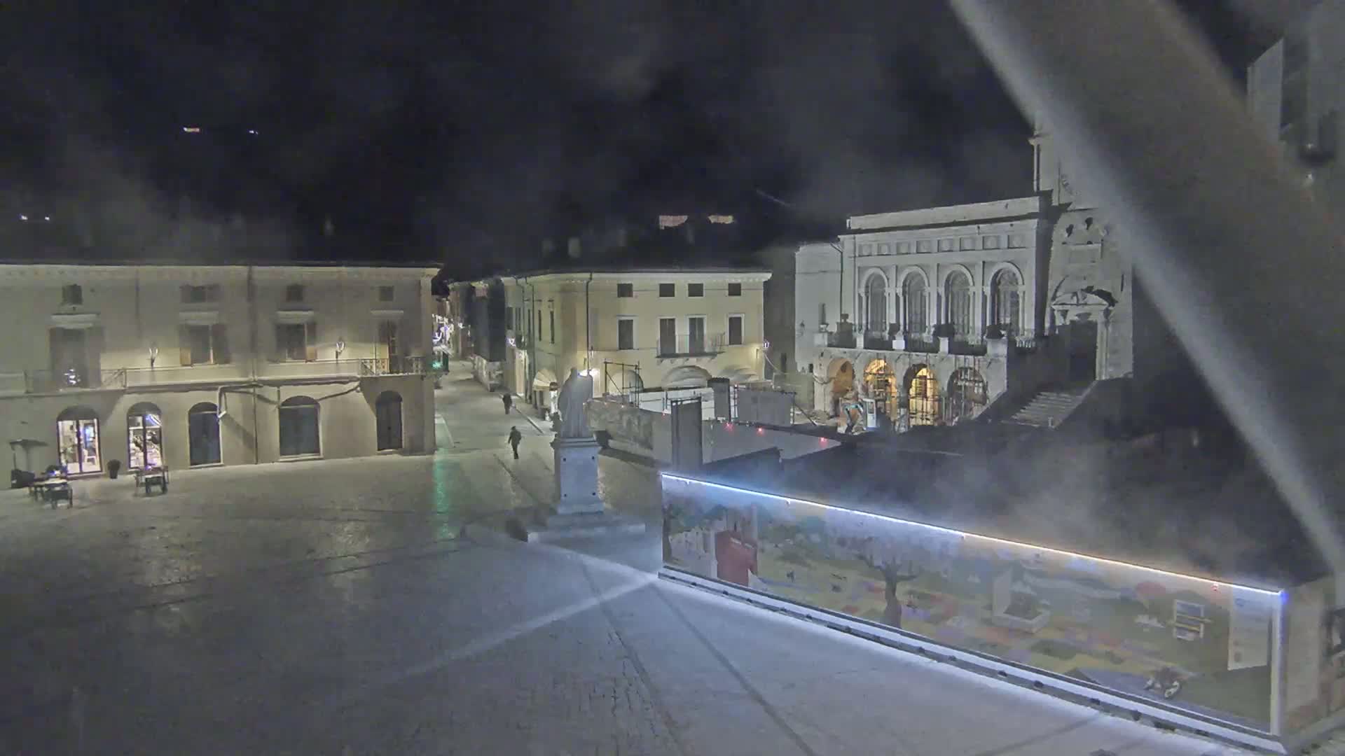A mostly empty town square at night, lightly covered in snow, is surrounded by buildings, one of which is under construction.