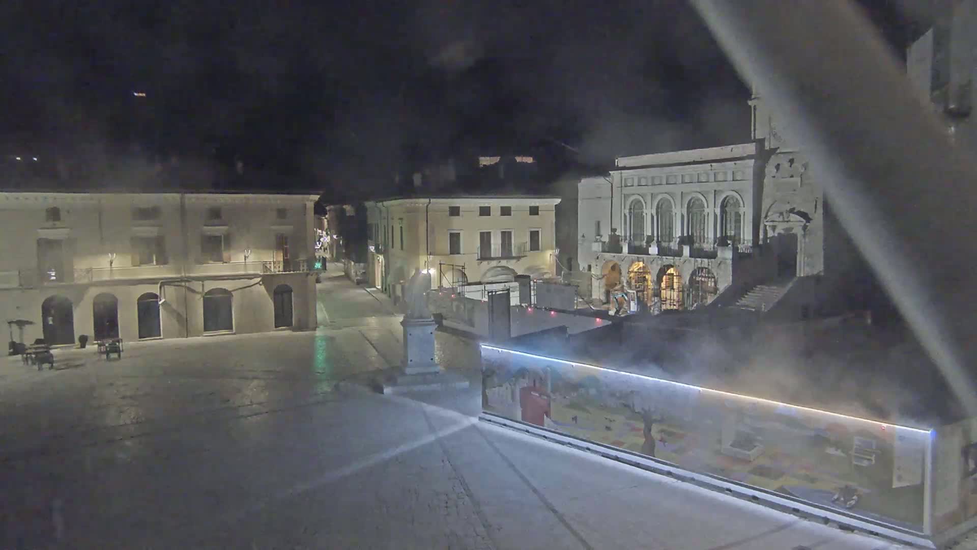 A snow-covered town square at night, with several buildings visible, one of which is large and ornate.