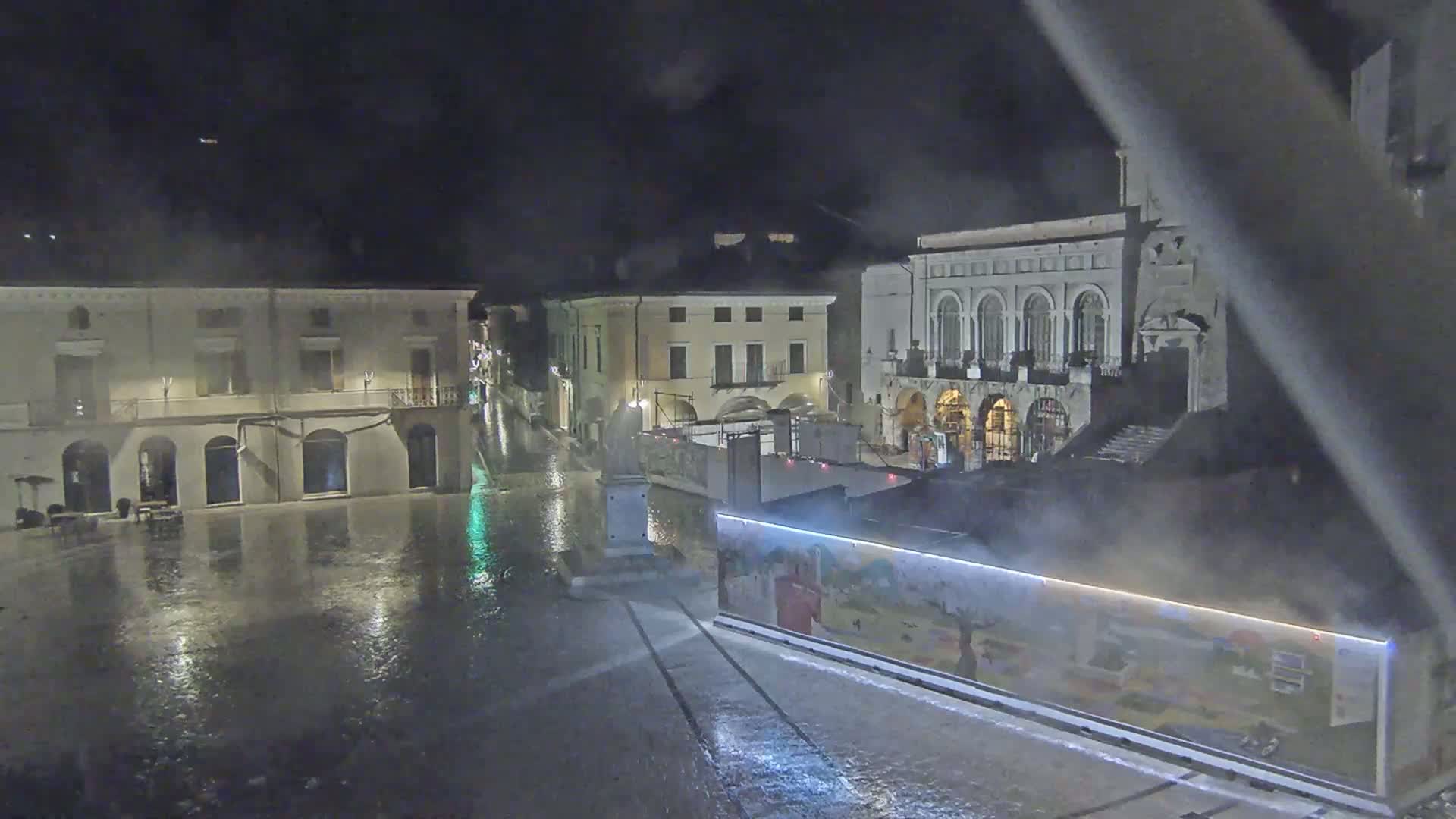 A rain-soaked town square at night, surrounded by buildings, with a large structure with artwork on its side in the foreground.