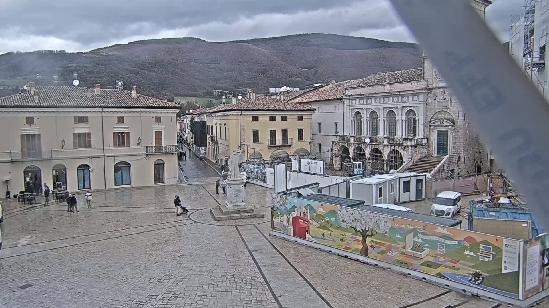 A town square with several buildings, a statue, and construction underway under an overcast sky.