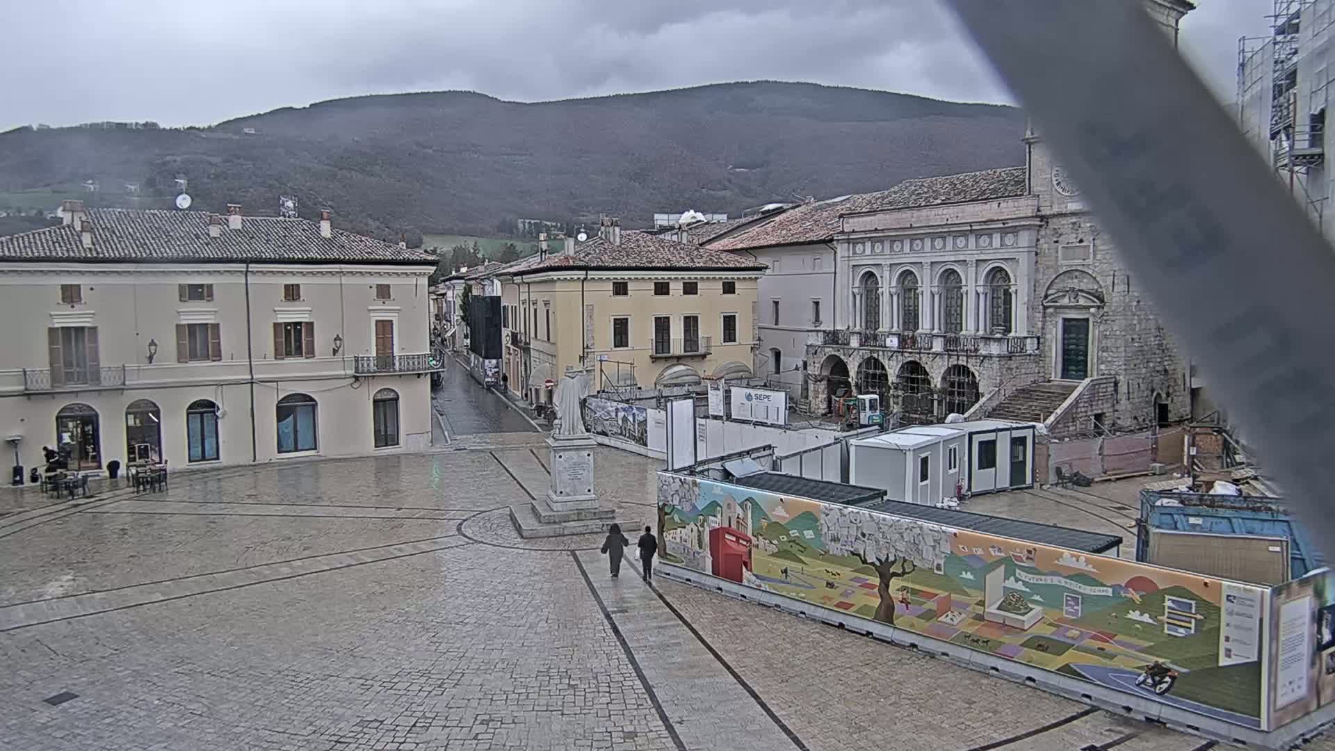 A town square under an overcast sky, featuring several buildings, construction barriers, and two pedestrians walking toward the center.