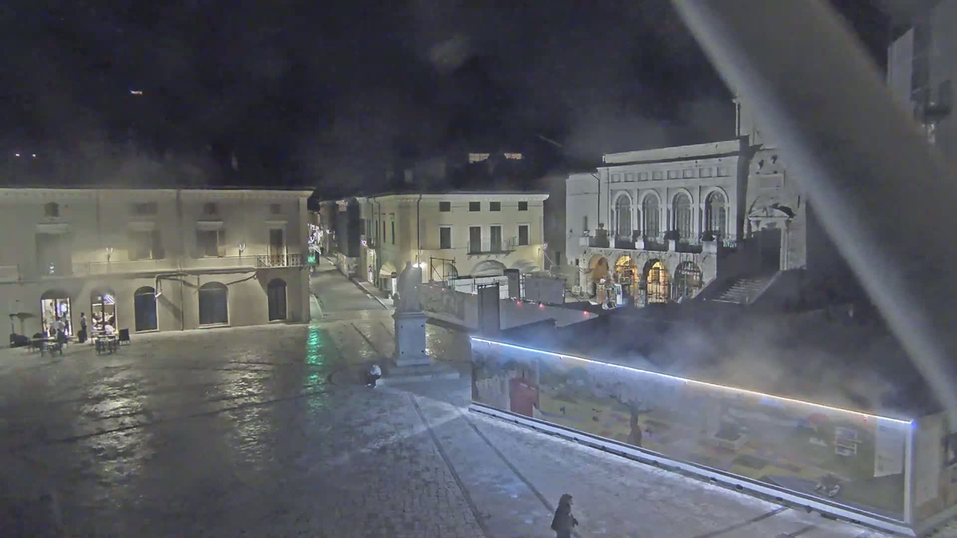 A nighttime view of a town square, partially covered in snow, with several buildings illuminated, and steam rising from a structure in the foreground.
