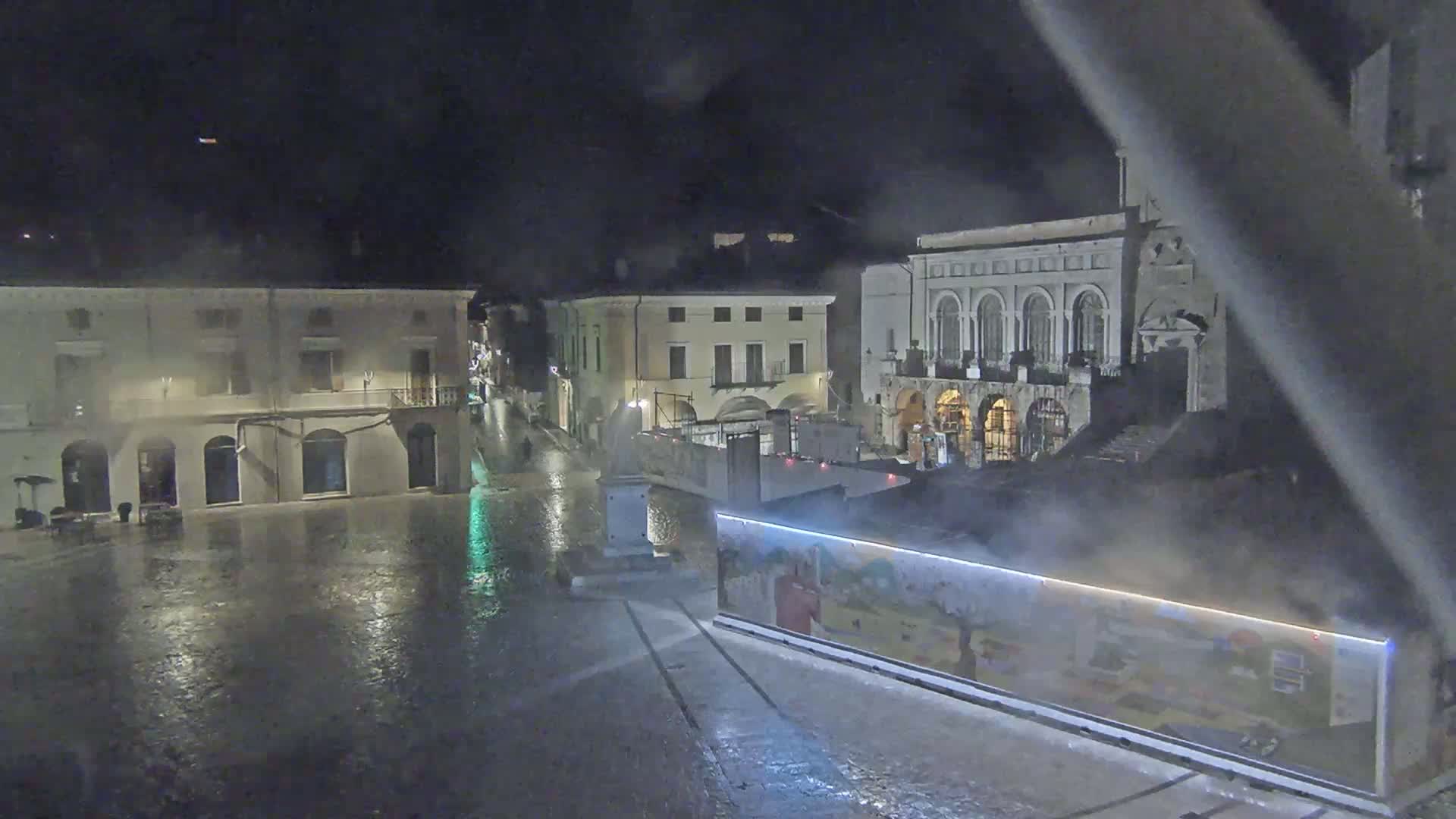 A rain-slicked town square at night, surrounded by buildings, with a large illustrated structure in the foreground.