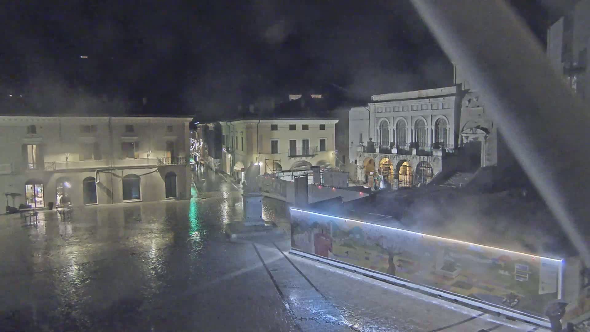 A rain-slicked town square at night, featuring several buildings, one of which is large and ornate, with a construction barrier and artwork along a section of the square.