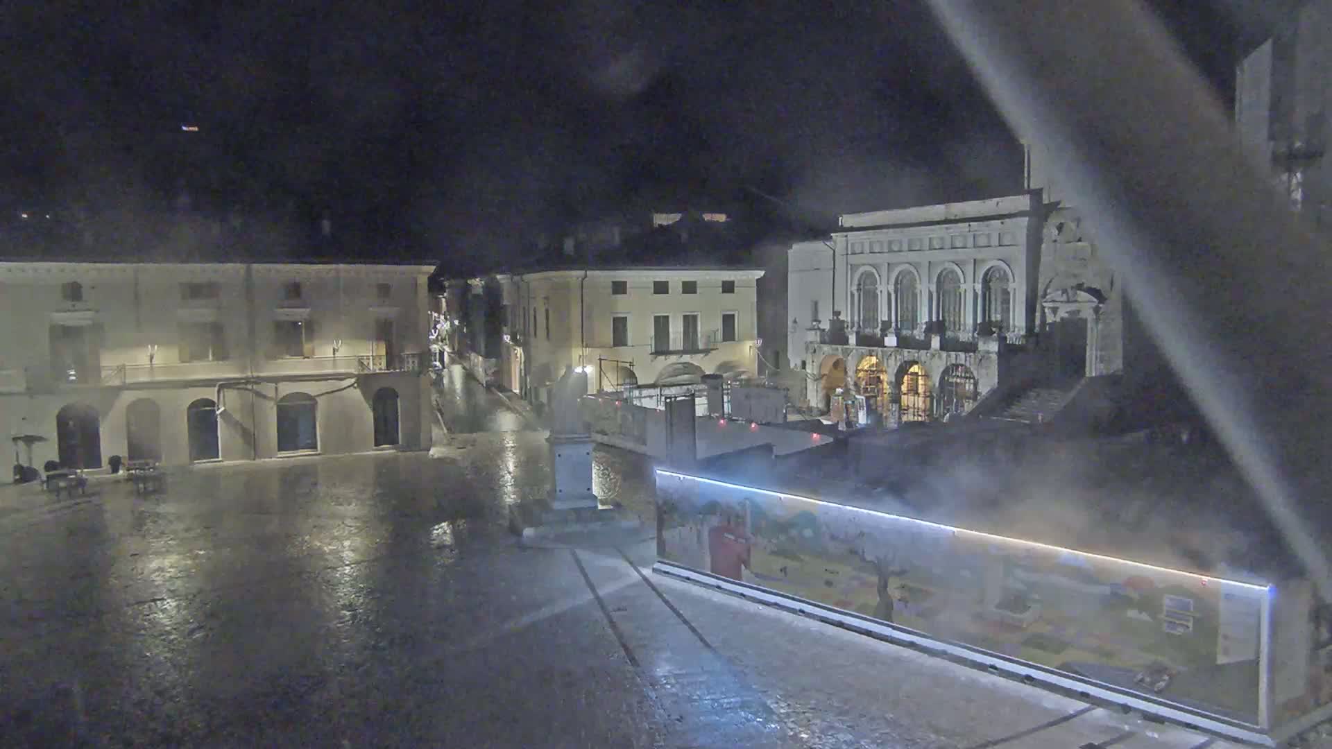 A rain-slicked town square at night, with several buildings visible, including one large building with arched entryways and a construction barrier in the foreground.