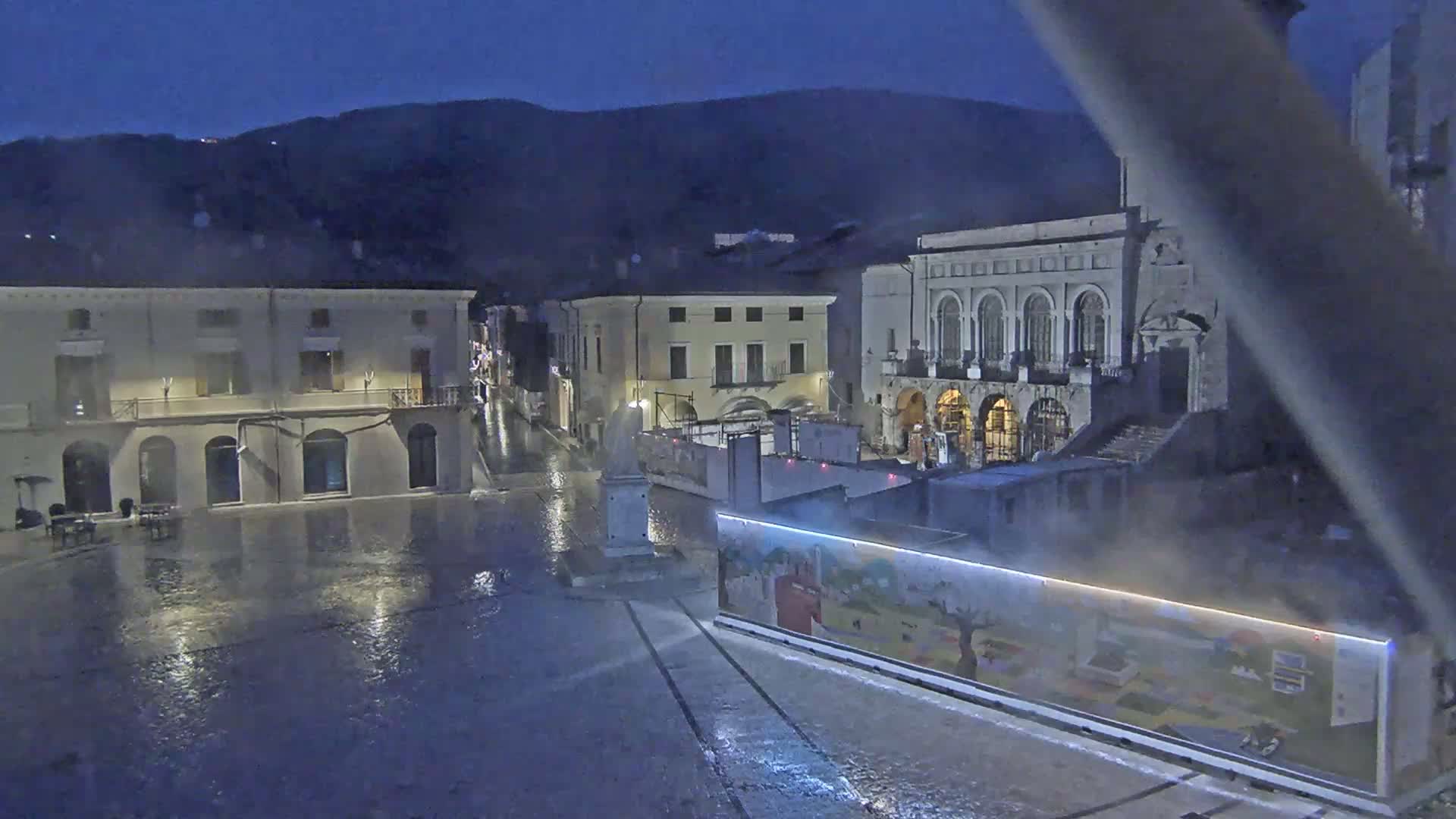 A rain-slicked town square at night, surrounded by old stone buildings under a dark sky, with mountains in the background.