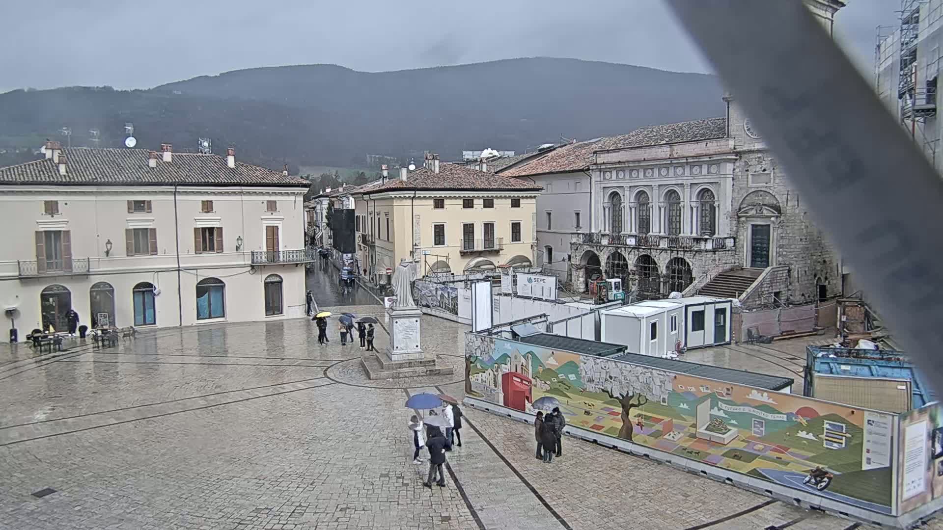 A rain-soaked town square, surrounded by historic buildings and construction, sits under a gray sky with mountains in the background.